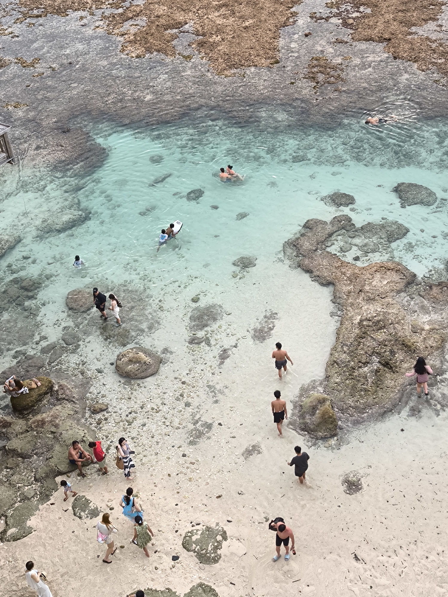 This aerial shot captures a vibrant beach scene in Uluwatu, Indonesia. The turquoise water reveals submerged rocks and sandy patches, where people are swimming, wading, and walking. Several individuals are swimming in the deeper areas of the water, while others stroll along the beach, interacting in small groups. Beachgoers are scattered across the sand, with some sitting on rocks and others wading in the shallow water. The coastline is characterized by rough, rocky formations, contrasting with the sandy beach and clear water.