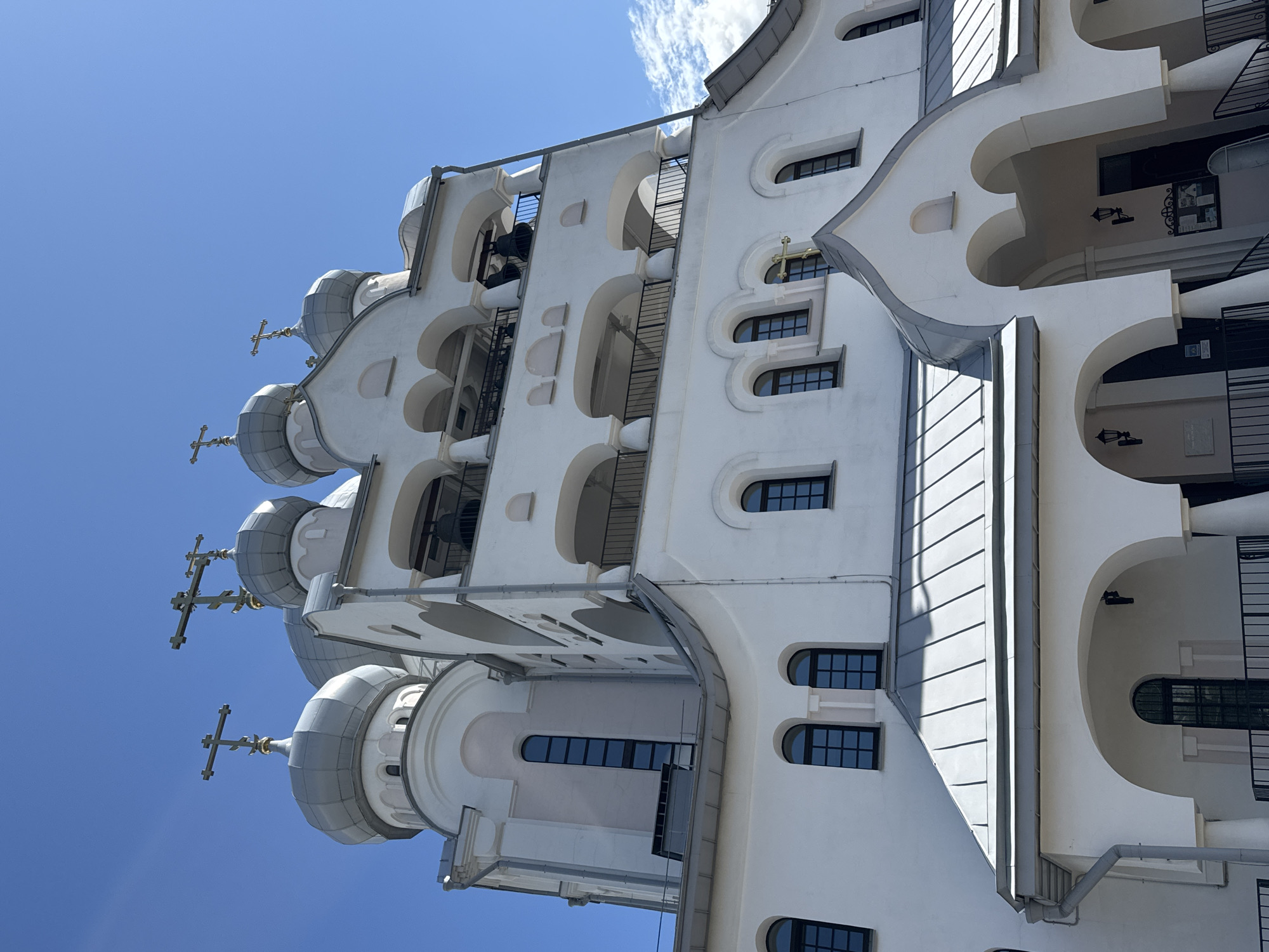The image shows a close-up, low-angle view of a white Orthodox church with multiple domes and crosses against a clear blue sky with a few wispy clouds. The architecture features arched windows, decorative moldings, and a bell tower with open arcades. The church is likely located in Saint Petersburg, Russia, a city known for its numerous religious buildings. There are no people visible in the image.