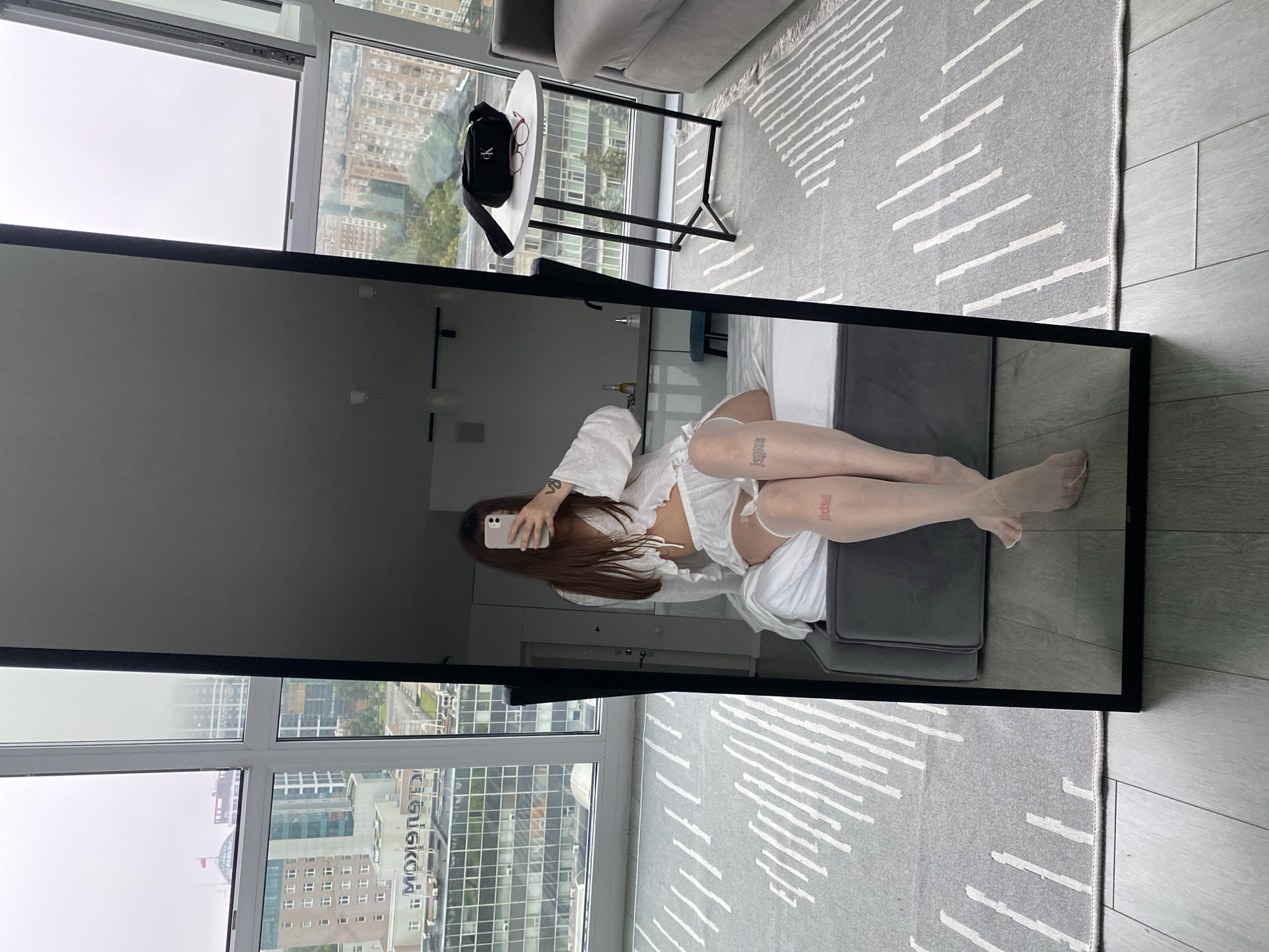A woman is taking a selfie in a full-length mirror in a modern apartment with a city view. She is sitting on the edge of a bed, wearing a white lace-trimmed top and matching shorts, with sheer stockings. Tattoos are visible on her legs. The apartment features a large window overlooking buildings in Izhevsk, Russia, a gray rug with a white geometric pattern, and wooden flooring. A small white table with a black frame holds a black fanny pack and glasses. The overall scene is bright and appears to be daytime.