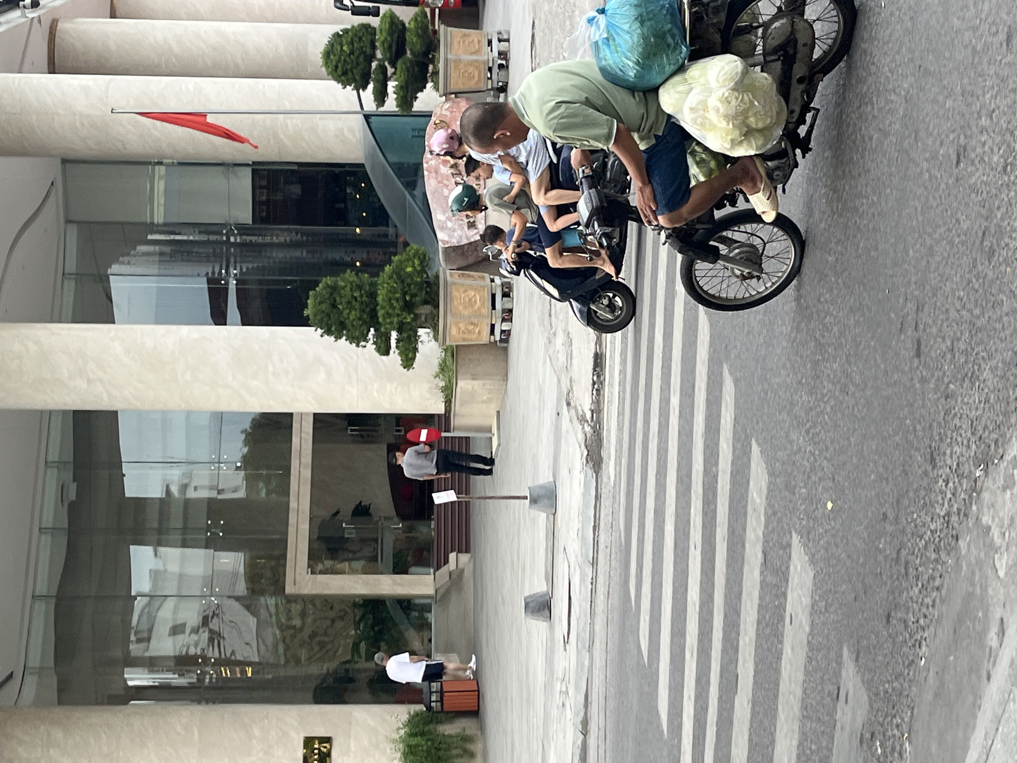 The image captures a street scene in Nha Trang, Vietnam, focusing on a pedestrian crossing and a modern building entrance. In the foreground, a man on a motorcycle is crossing a pedestrian street, carrying large bags of produce. He is wearing a light green short-sleeved shirt and blue shorts, with white sandals. Behind him, another motorcycle carries four people: a man in a green helmet, a woman with a pink helmet, and two children. The building in the background features large glass windows reflecting the surroundings and tall, cream-colored pillars. A red flag flies from a pole near the entrance. Two men are visible near the building's entrance: one in a white t-shirt and shorts, and another in a gray t-shirt and dark pants. The environment suggests a commercial or hotel area with well-maintained landscaping.