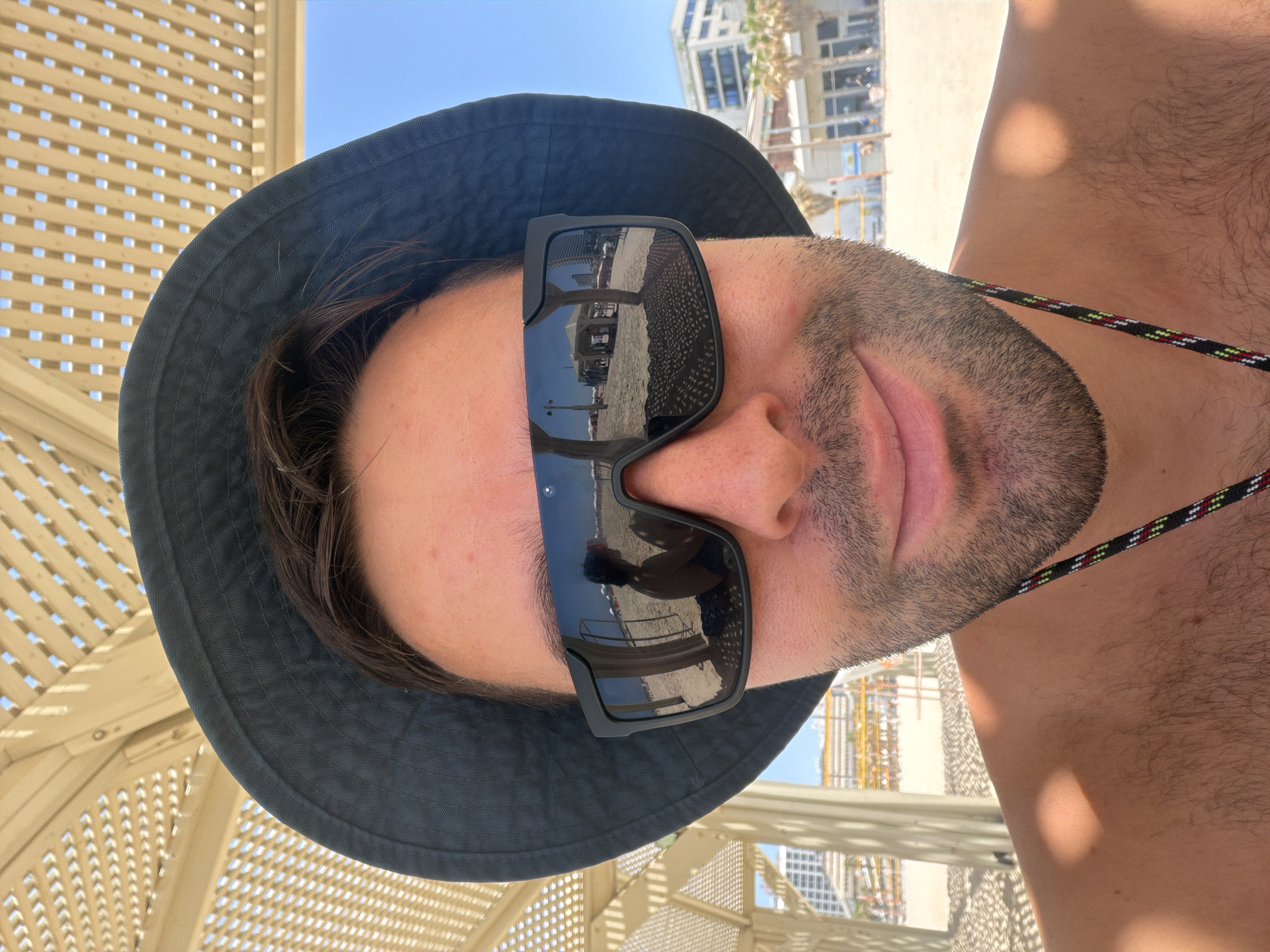 A man is taking a selfie at a beach in Tel Aviv, Israel. He is wearing a dark bucket hat and large black sunglasses, which reflect a scene of the beach with people sitting under umbrellas. The man has a light beard and is shirtless, with his shoulder and chest visible. The background shows a sandy beach with rows of beach umbrellas, palm trees, and buildings in the distance. The sky is clear and blue. The man is partially shaded by a wooden slatted structure overhead.