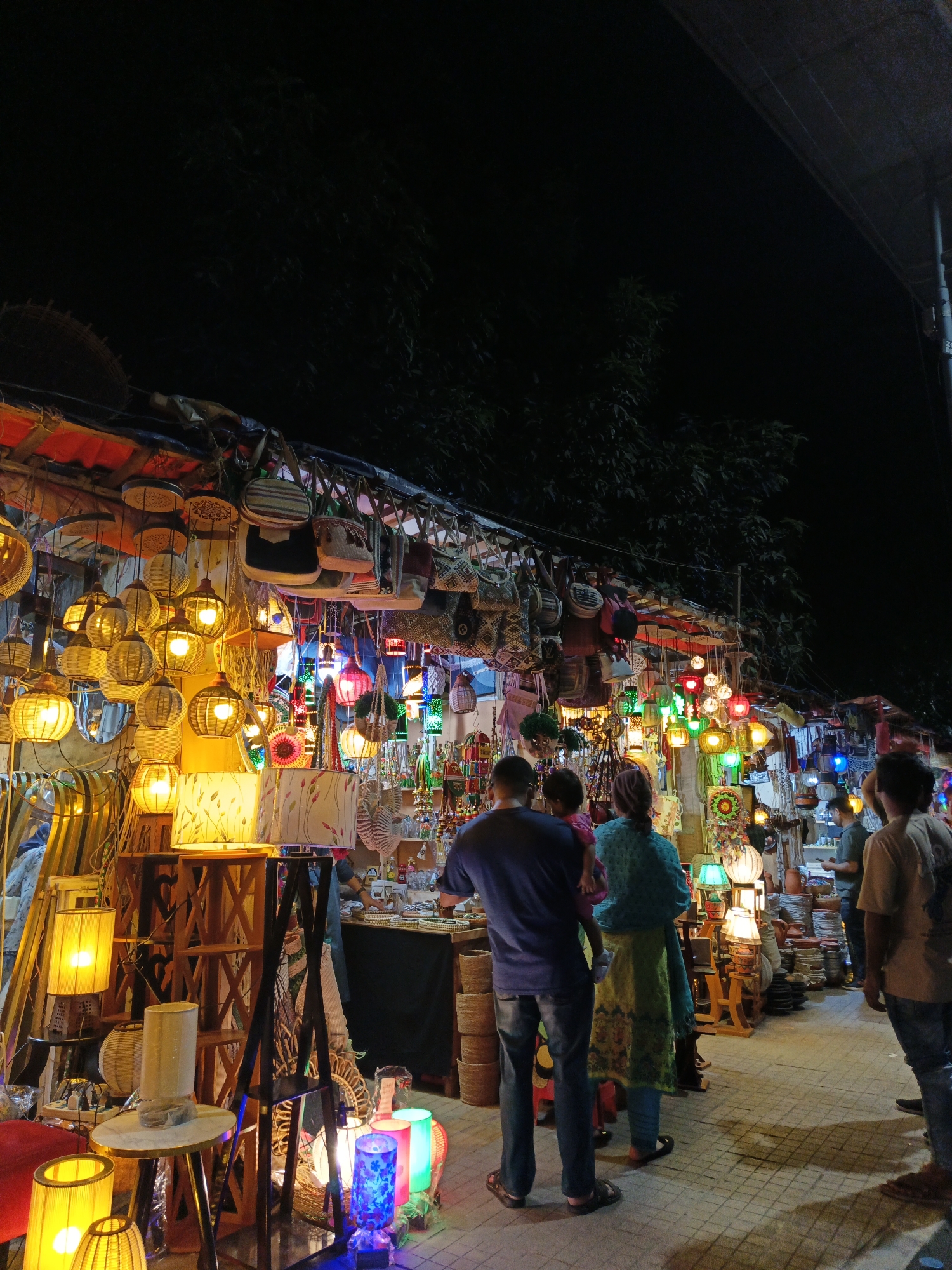 This is a night-time photograph of a street market in Dhaka, Bangladesh, likely a craft bazaar. The scene is illuminated by numerous colorful lights, predominantly yellow, emanating from hanging lamps, table lamps, and decorative lanterns. A variety of handcrafted items are displayed for sale, including woven bags hanging from the stalls, intricately designed lampshades, and pottery.
In the foreground, several people are seen browsing the stalls. A man in a dark blue shirt and jeans stands facing away from the camera, holding a child in a pink garment. To his right, a woman in a teal blue printed kurti and a green patterned skirt stands with her back to the viewer, also facing the market displays. Further to the right, another person in a light brown t-shirt and jeans is partially visible, looking towards the stalls. In the mid-ground, more people are present, some interacting with vendors.
The market appears to be set up under a temporary shelter with a red roof structure visible above the stalls. The ground is paved with light-colored tiles forming a grid pattern. The overall atmosphere is vibrant and busy, characteristic of a local night market.