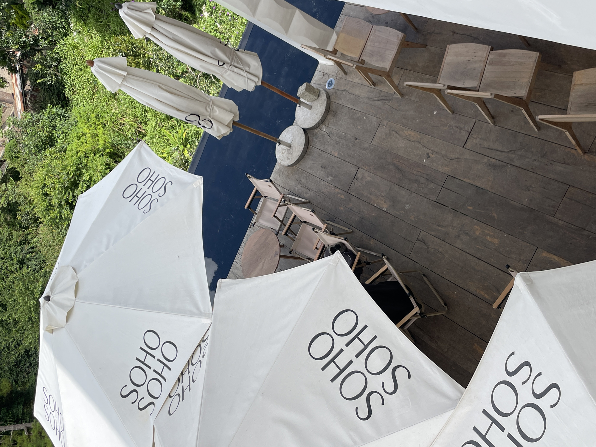 The image depicts an outdoor seating area at the SoHo SoHo cafe in Da Lat, Vietnam. The scene is set on a wooden deck overlooking lush greenery. Three white umbrellas are prominently featured, two of which are open and display the "SOHO SOHO" logo in black lettering. One of these open umbrellas is partially obscuring another, closed umbrella. Behind these, two more closed, beige umbrellas stand upright in concrete bases. Scattered across the wooden deck are several chairs, some made of light-colored wood and others appearing to be folding chairs with fabric seats. A round wooden table is visible near the closed umbrellas. The environment is natural and verdant, with dense foliage forming a backdrop. The lighting suggests it is daytime.