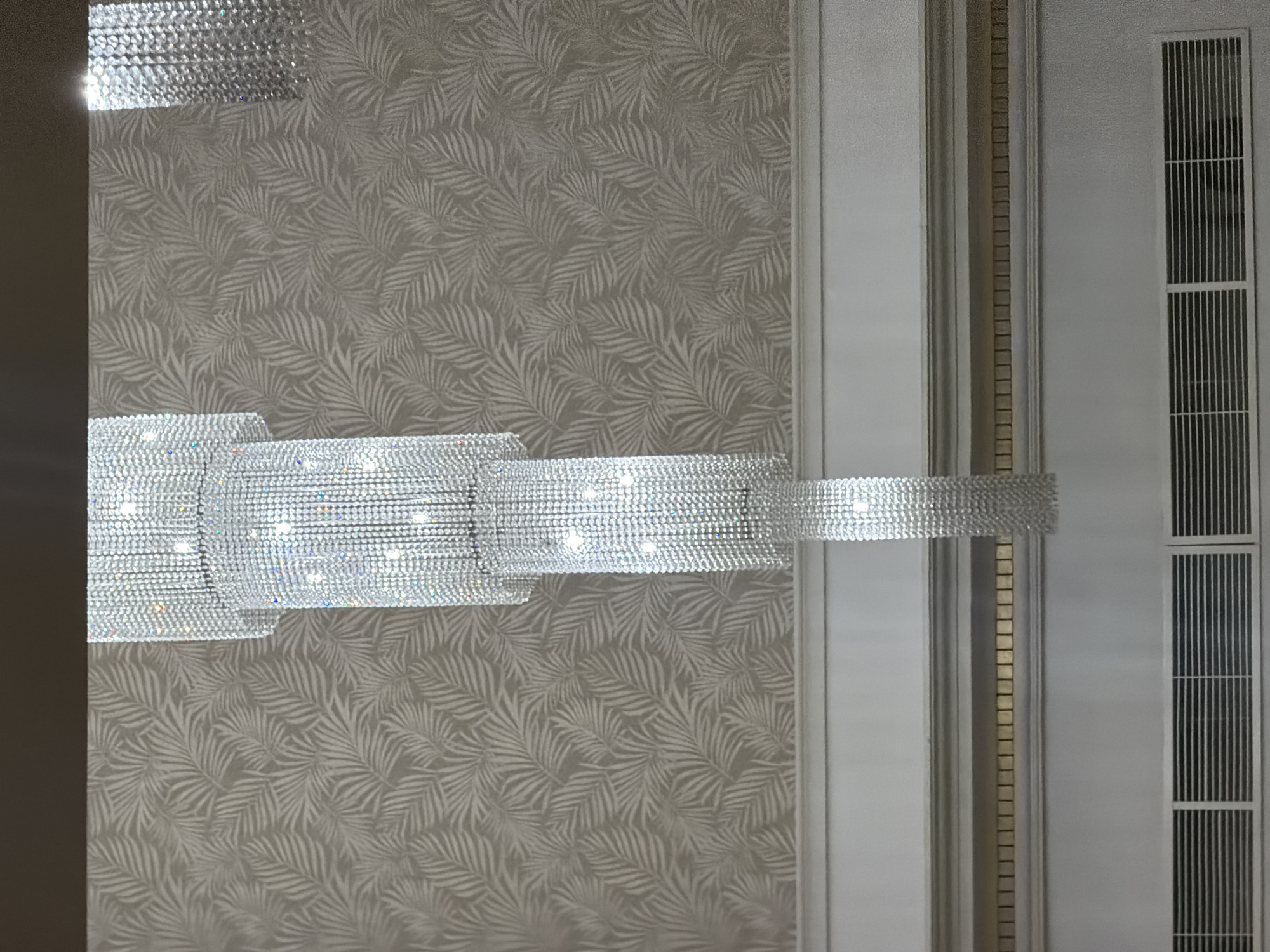 This image captures a detail of an elegant interior setting, focusing on a large, multi-tiered chandelier and decorative wall elements. The prominent subject is a horizontal segment of a shimmering chandelier, heavily adorned with numerous small, clear crystals or beads that reflect light intensely, creating bright sparkles and streaks. The chandelier appears to be illuminated from within, casting a soft glow.
Behind the chandelier, the wall is covered with wallpaper featuring a repeating pattern of stylized palm leaves in a lighter shade against a soft, grayish-beige background. The pattern adds a subtle texture and organic motif to the luxurious environment.
To the right side of the frame, there are architectural details. A light-colored column or panel is visible, next to which runs a narrow, vertical strip adorned with small, square, tile-like elements, possibly in a golden or light brown hue. Further to the right, a white panel features two rectangular grilles, likely air vents or decorative screens, composed of thin, dark vertical slats.
No people are present in the image, and no specific activity or event is taking place, presenting a static view of the decor. The indoor lighting from the chandelier suggests either nighttime or an interior space where artificial illumination is primary. The overall impression conveyed is one of sophistication and opulence. No visible text is present.