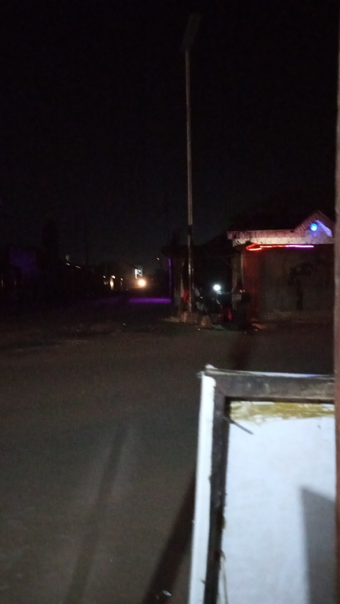 Night scene in Sabon Tasha, Nigeria. A dark road extends into the distance with the faint glow of vehicle headlights visible. A building with red and blue decorative lights is situated on the right. A tall streetlight is on the left. A white door frame is in the foreground. There are no people visible.