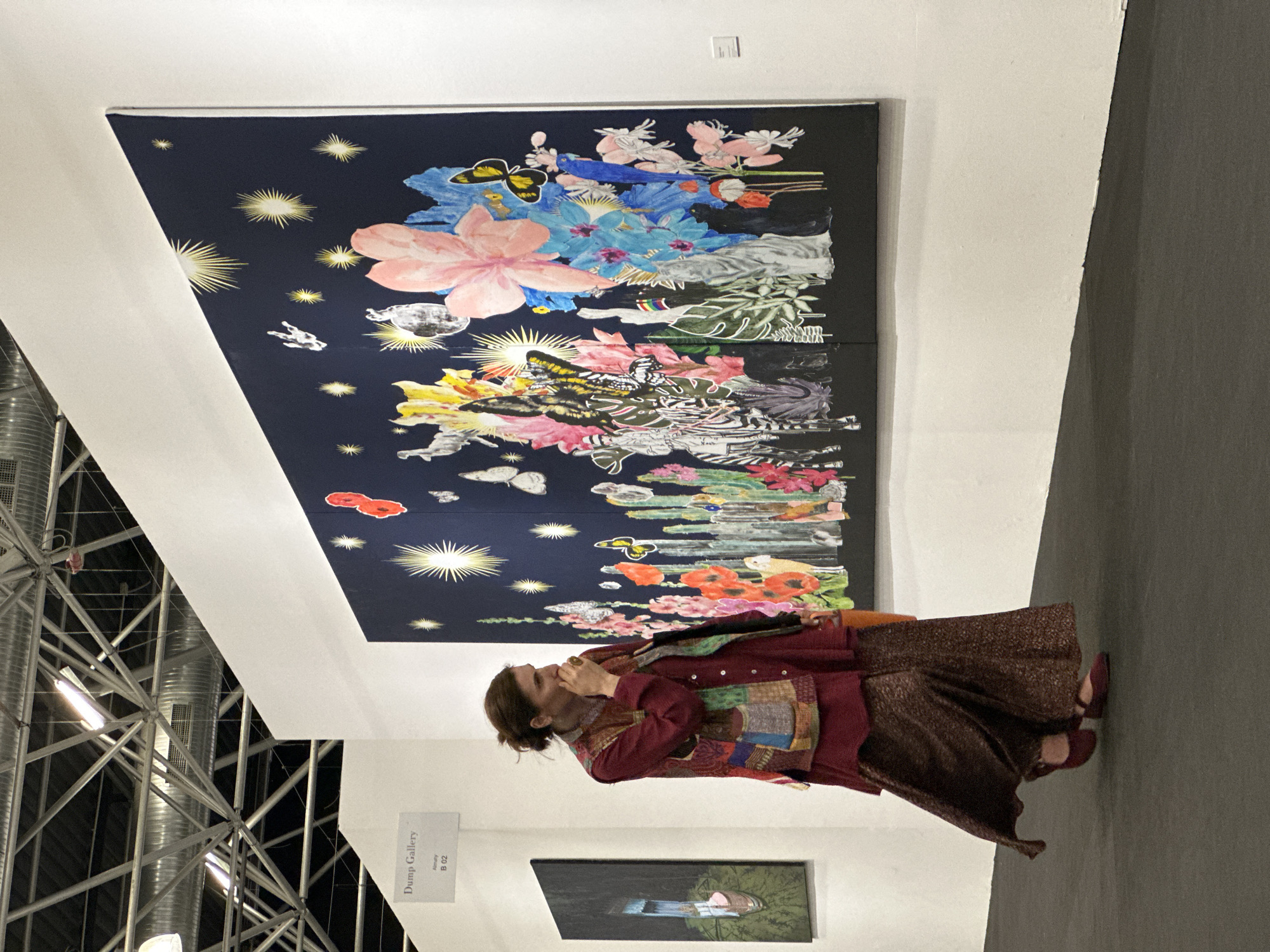 A medium indoor shot features a woman standing in front of a large painting at what appears to be an art gallery, possibly in Tbilisi, Georgia. The painting is primarily blue, with floral designs and star-like patterns, and is placed high on the wall. The woman has her hair pulled back and is dressed in a burgundy jacket and patterned skirt. She is facing the painting and appears to be holding something near her mouth. Below the painting, there is another artwork on display. A sign with the text "Dump Gallery" is visible on the wall. The gallery is inside what appears to be a large exposition hall with industrial metallic structures.