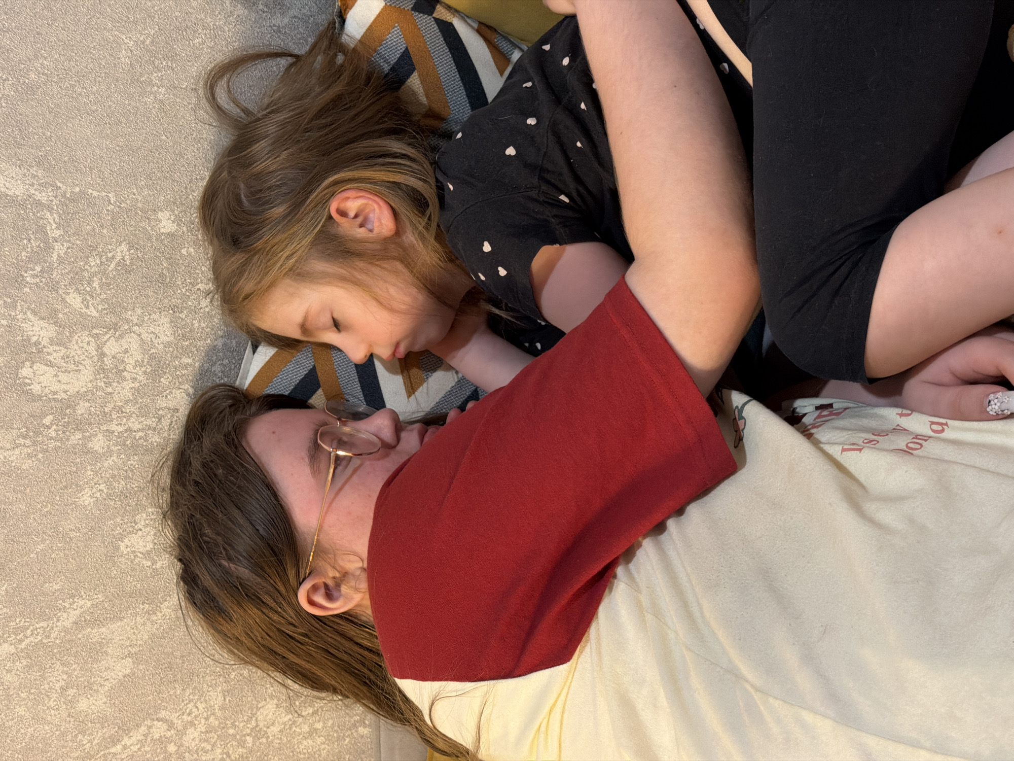 In the city of Perm, Russia, two young girls are lying down, embracing each other and facing each other. The girl on the left, wearing a red and cream shirt and glasses, has her eyes open and is looking at the girl on the right. The girl on the right, wearing a black shirt with white hearts, has her eyes closed and appears to be sleeping or resting. They are positioned on a couch with a patterned pillow behind them, and a textured wall is visible in the background. The overall scene suggests a moment of affection and comfort between siblings or close friends.