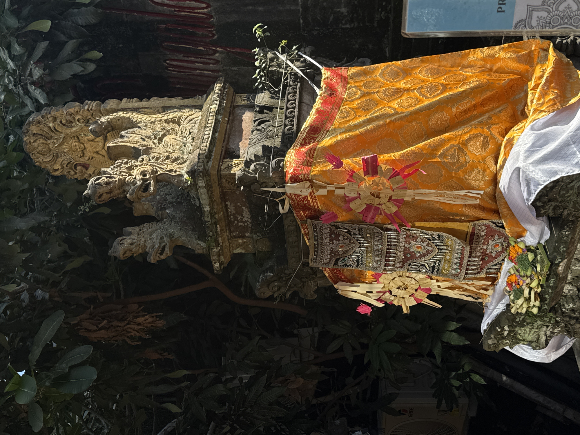 In Ubud District, Indonesia, a stone altar, adorned with an elaborate yellow and red fabric draped over its lower section, is the focal point. The altar is decorated with intricate carvings and features religious offerings, including flowers and what appears to be fruit. To the left, an air conditioning unit is partially visible. The background is dominated by lush green foliage and a stone wall with red markings. The lighting suggests it is daytime, with sunlight casting shadows.