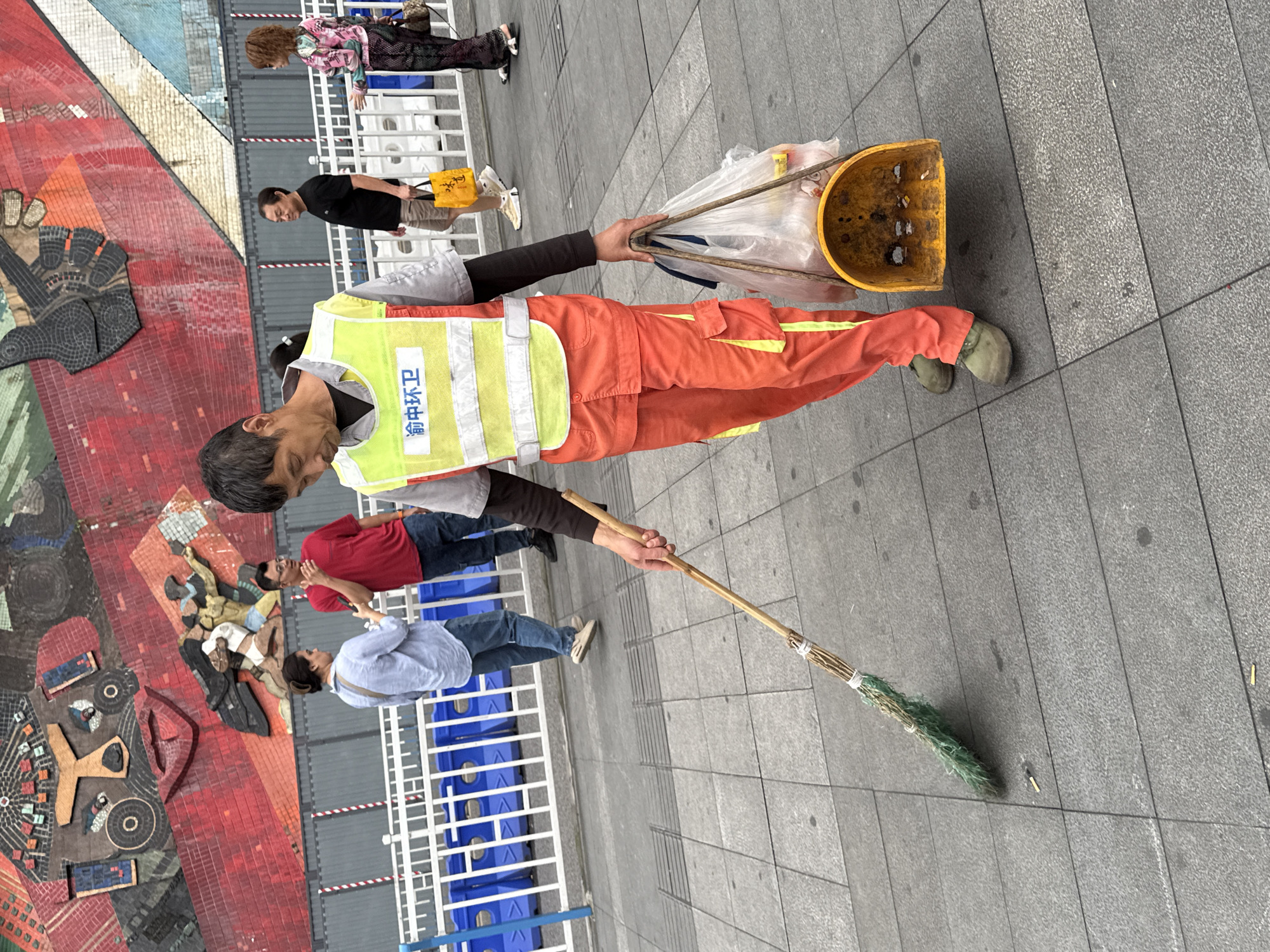 A street sweeper in orange pants and a yellow safety vest sweeps a sidewalk in Yuzhong District, China. The person sweeps with a straw broom in the right hand and carries a yellow scoop full of garbage in the left hand. Several pedestrians walk in the background. A colorful mosaic mural appears on the left, and a metal barricade borders the walkway. The sidewalk consists of gray pavers.