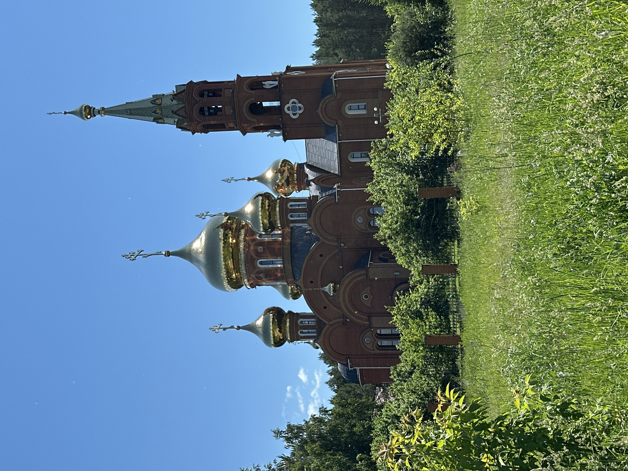 The image depicts a brick Orthodox church with golden domes against a clear blue sky. The church features a prominent bell tower with arched openings and a tall spire topped with a cross. The building is surrounded by lush green trees and foliage, with a tall grassy field in the foreground. A fence made of brick posts and metal bars separates the church grounds from the field. The scene appears to be a serene, outdoor environment. The church is the Church of the Nativity of Christ, located in Chaikovsky, Russia.