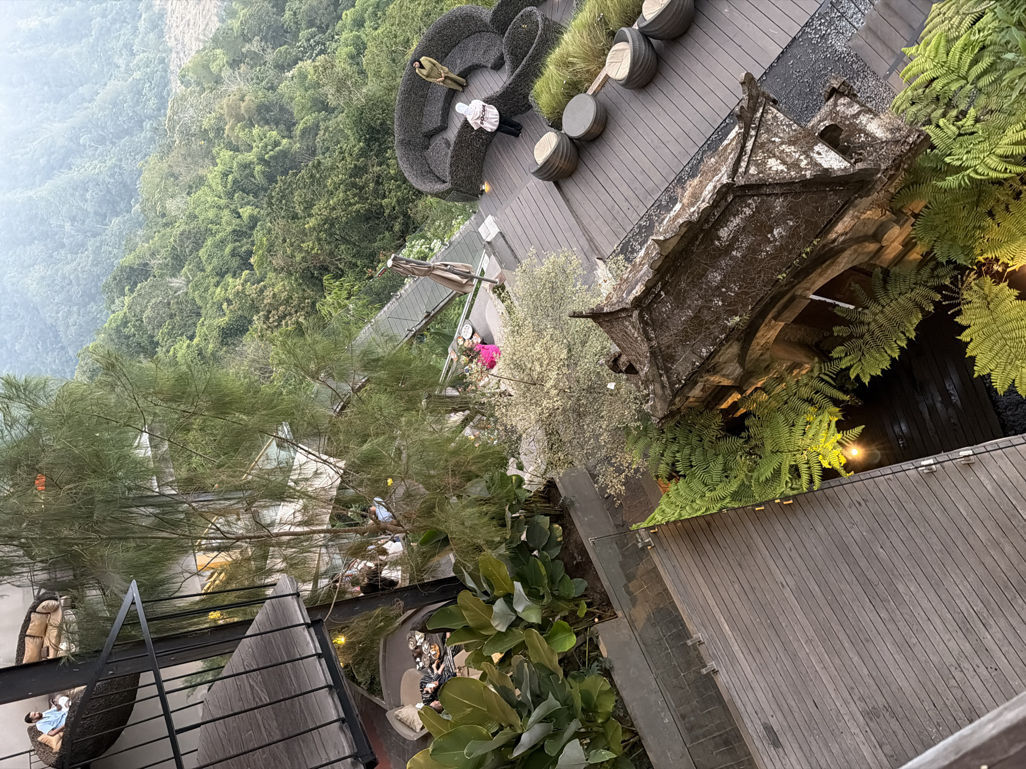 An elevated, high-angle shot captures a portion of a multi-level resort complex in Kintamani, Indonesia, with lush, misty mountains dominating the background. The foreground features a weathered wooden deck with fern plants and a portion of an old, stone-tiled roof structure.
On the mid-level, a dark, woven circular sofa-like seating area is visible, with two individuals. One person, wearing a light-colored hijab and hoodie with "SOCIAL" text, stands facing away. Another person, wearing a tan outfit and a headscarf, stands facing the first individual. Several round, woven stools and potted grass are arranged on the wooden deck. Adjacent to this area, a person in a pink outfit sits on a low table with food.
Further down, a large planter box holds lush, broad-leafed green plants. A metal railing encloses a wooden balcony area, where a man wearing a light blue shirt sits on a woven chair, holding a phone. In the background, other levels of the resort are visible, with lights illuminating indoor spaces and balconies. A long, elevated walkway or bridge crosses over a lower deck. The overall scene suggests a tranquil, scenic resort with natural elements integrated into its architecture.