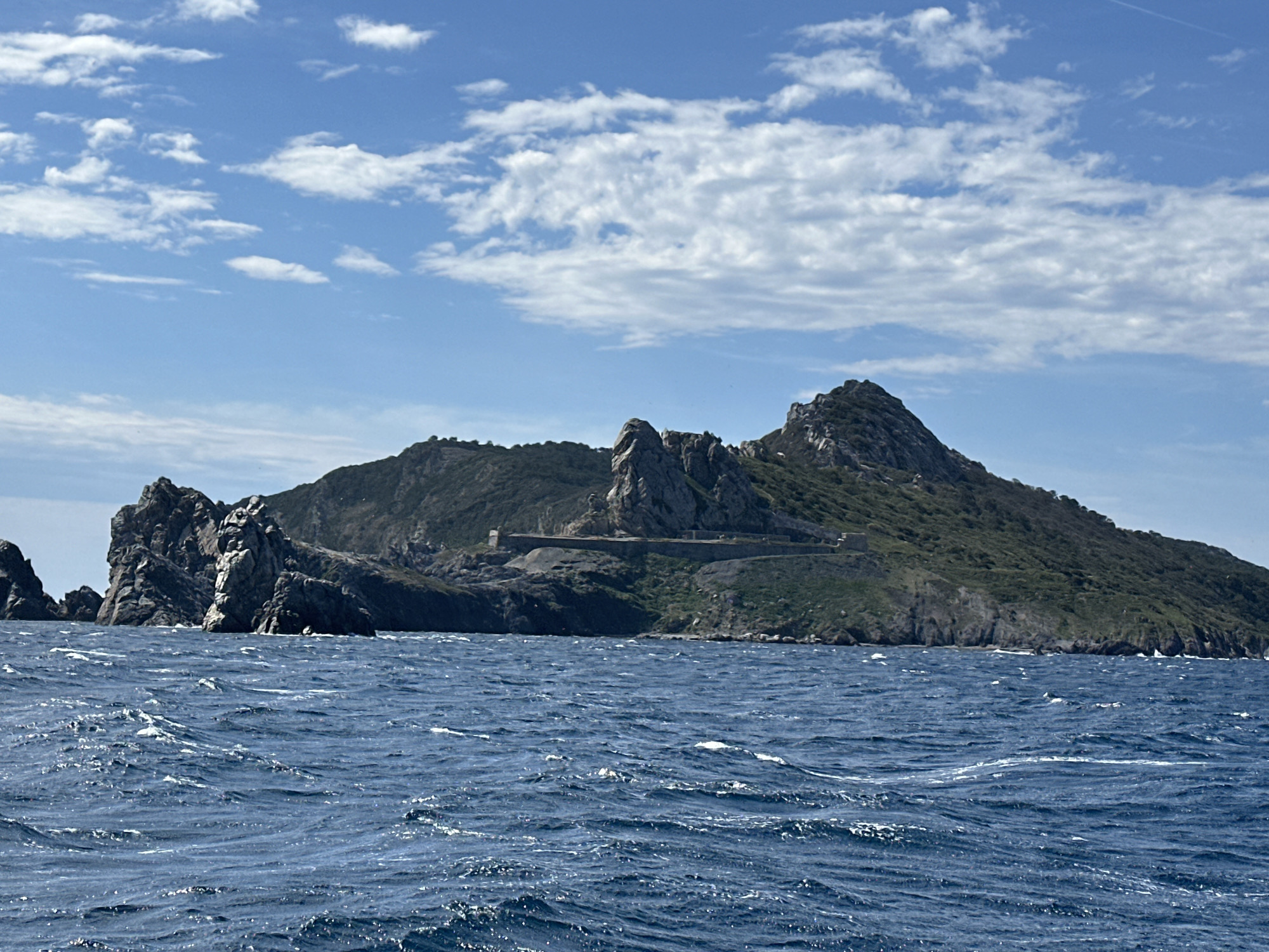 The image shows a view of the island of Porquerolles, part of the Îles d'Hyères, off the coast of Hyères, France. A rugged, green, and rocky island rises from the deep blue, choppy sea. The island is characterized by steep cliffs, dense vegetation, and prominent rock formations. Along the mid-section of the island, a long, weathered stone wall or structure is visible, suggesting some form of historical construction. The sky above is a bright blue, dotted with scattered white clouds. The foreground is dominated by the textured surface of the ocean, with waves breaking and creating white foam. No people or specific man-made objects, beyond the stone structure on the island, are discernible. The scene depicts a natural coastal landscape under a partly cloudy sky.