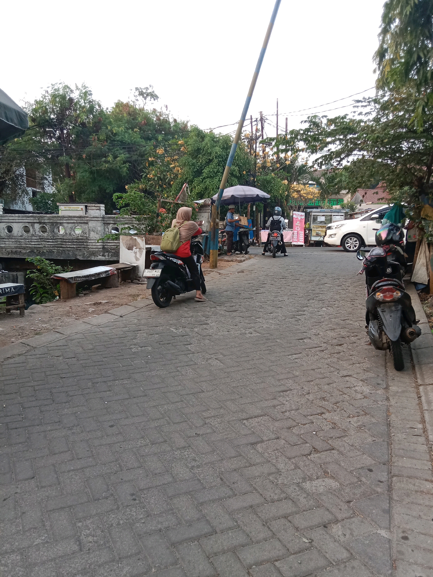 The image depicts a street scene in South Tangerang, Indonesia, characterized by a brick-paved road lined with trees and buildings. A person wearing a hijab and a backpack rides a black motorcycle on the right side of the road. Several other motorcycles and a white car are visible further down the street. A white, decorative bridge with circular openings spans a small waterway on the left side of the image. Overhead, a striped pole extends across the scene, suggesting it might be part of a barrier or gate. Various food stalls and shops are present along the roadside, indicating a commercial area. The sky is overcast, suggesting a cloudy day.