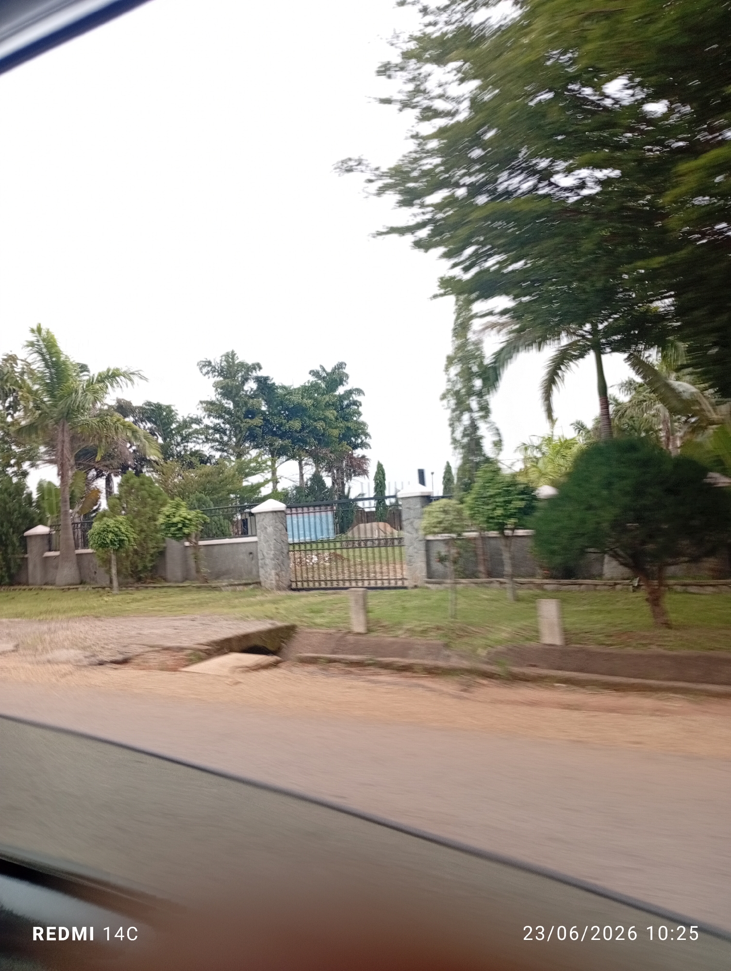 The image shows a view of a property entrance in Du, Nigeria. The entrance features a grey stone pillar with a metal gate. Behind the gate, there are various trees and shrubs, including palm trees and manicured bushes. The property appears to be well-maintained with a paved driveway leading up to the entrance. The sky is overcast, and the overall lighting suggests it is daytime. A portion of a vehicle's interior, including the dashboard and window, is visible in the foreground, indicating the photo was taken from inside a car. The timestamp in the lower right corner reads "23/06/2026 10:25".