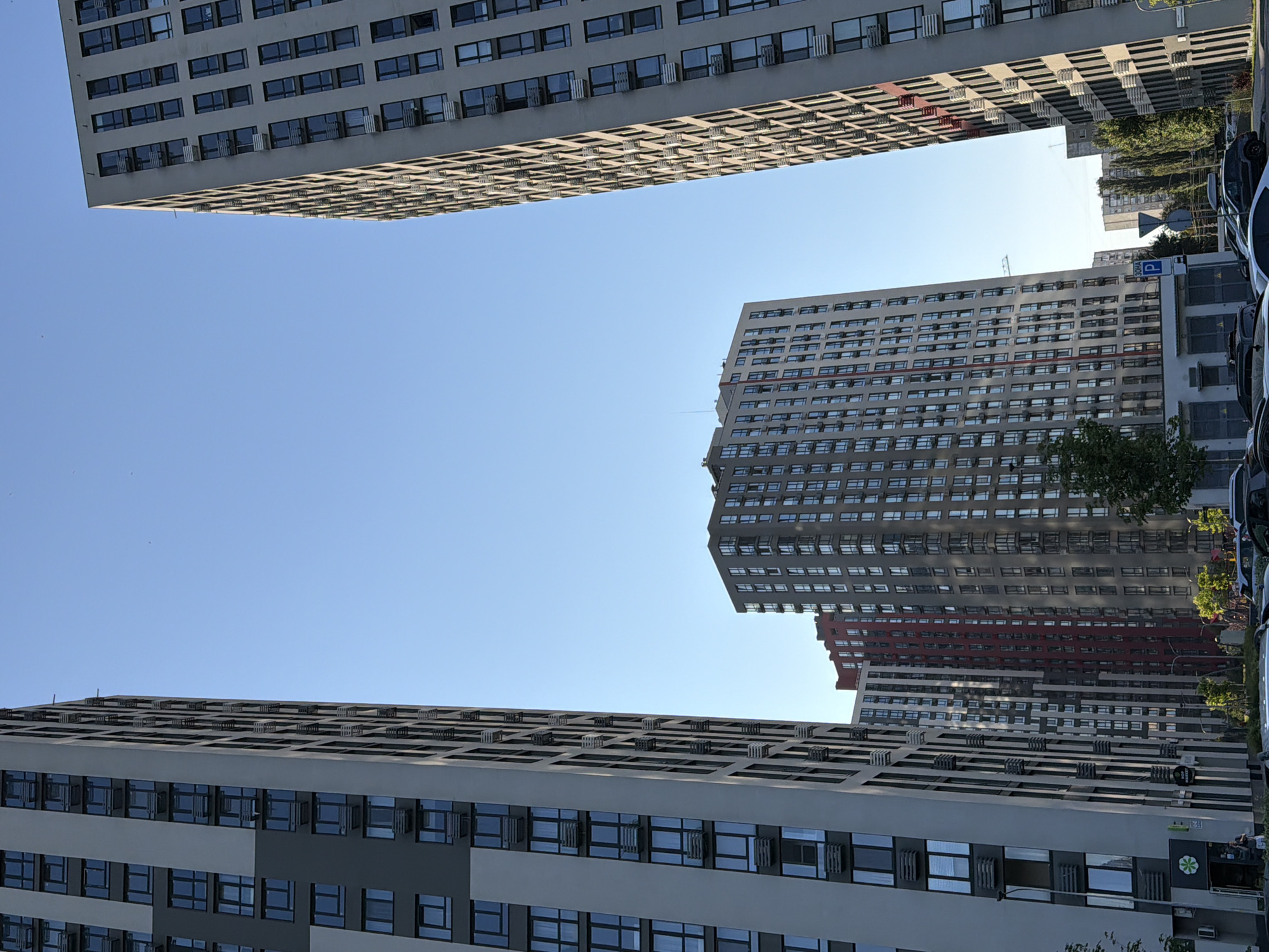 This is a photograph of a modern residential complex in Kyiv, Ukraine, captured from a low angle looking upwards at the tall buildings against a clear blue sky. The buildings are contemporary high-rise apartment blocks with numerous windows and air conditioning units visible on their facades. In the foreground, on the left, is a building with a grey and white exterior, featuring vertical divisions and rows of identical windows. To its right, a taller, narrower building with a similar design but a predominantly grey facade dominates the right side of the frame. In the middle ground, a cluster of buildings of varying heights and colors, including a prominent red-toned structure, are visible. The scene appears to be a newly developed urban area, possibly a residential microdistrict, characterized by the density of high-rise construction. The presence of trees and some parked cars at the base of the buildings suggests an active urban environment. The lighting indicates it is daytime, with sunlight casting shadows. There are a few individuals visible near the entrance of the building on the left.