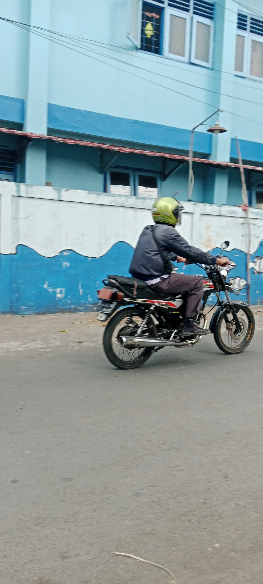A person wearing a green helmet and a dark jacket rides a motorcycle on an asphalt road. The motorcycle is black and red, with a silver exhaust pipe. The person is wearing dark pants and black shoes. In the background, there is a light blue building with white trim and windows. A blue and white mural runs along a wall in front of the building. The scene appears to be in an urban environment in Purwomartani, Indonesia, indicated by the building architecture and the style of the motorcycle and mural.