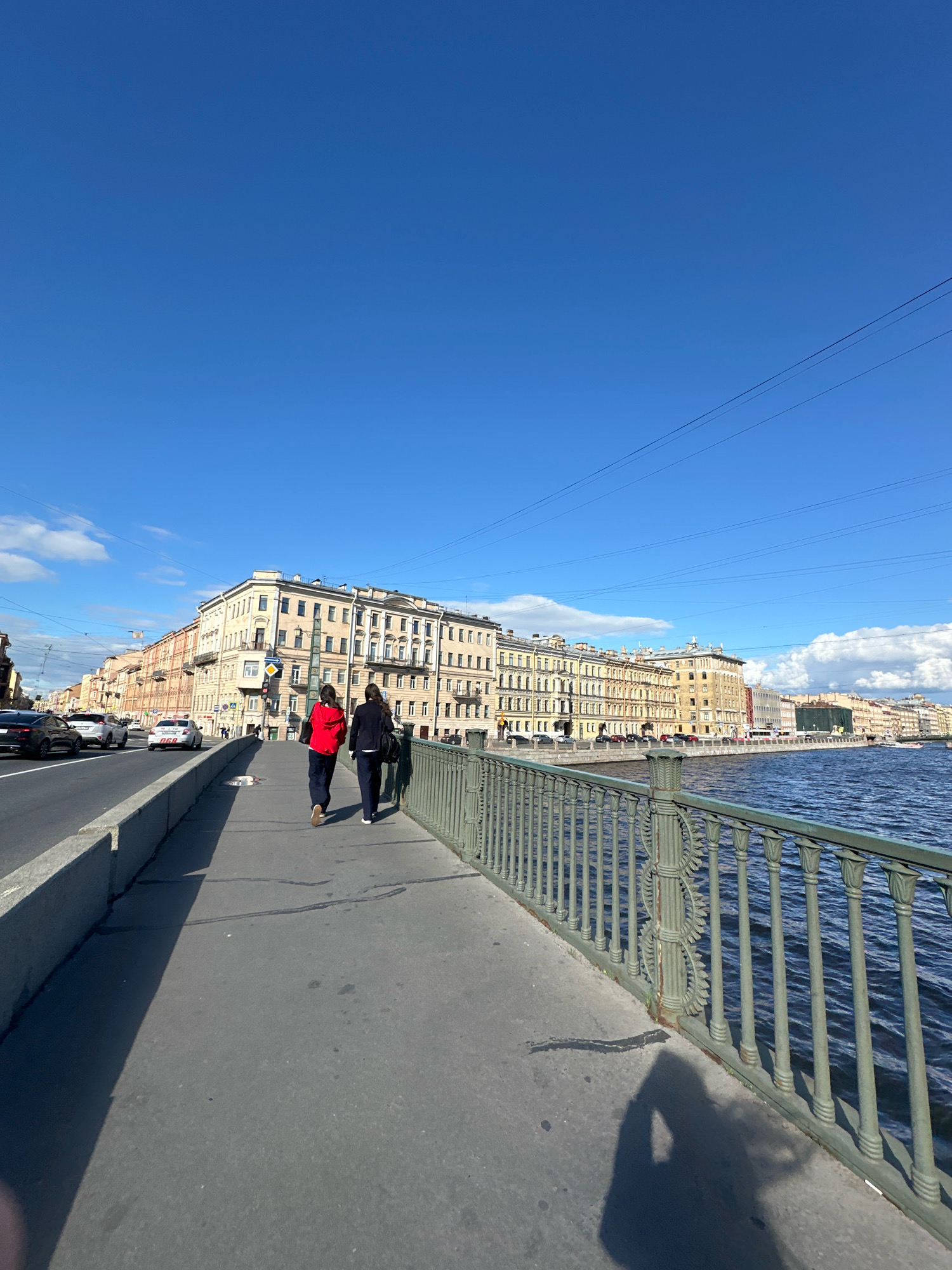 This is a photograph taken from a pedestrian walkway on a bridge over a body of water in Saint Petersburg, Russia. The viewpoint is looking towards a long row of historic, multi-story buildings lining the opposite bank. The architecture suggests the buildings are typical of 19th-century European urban construction, with numerous windows and decorative elements.
In the foreground, two individuals are walking away from the camera along the walkway. The person on the left is wearing a bright red jacket and dark pants, while the person on the right is wearing a dark suit jacket and dark pants. A dark green metal railing with decorative detailing runs along the edge of the walkway, separating it from the water. The water appears to be a river or canal, with visible ripples and a dark blue color.
To the left of the walkway, there is a road with several cars driving on it. The road is separated from the pedestrian walkway by a concrete barrier. The sky above is a clear, bright blue with a few scattered white clouds. The sunlight casts long shadows on the walkway, indicating it is likely late afternoon or early morning. The overall scene depicts a typical urban environment with a blend of historical architecture, modern infrastructure, and everyday human activity.