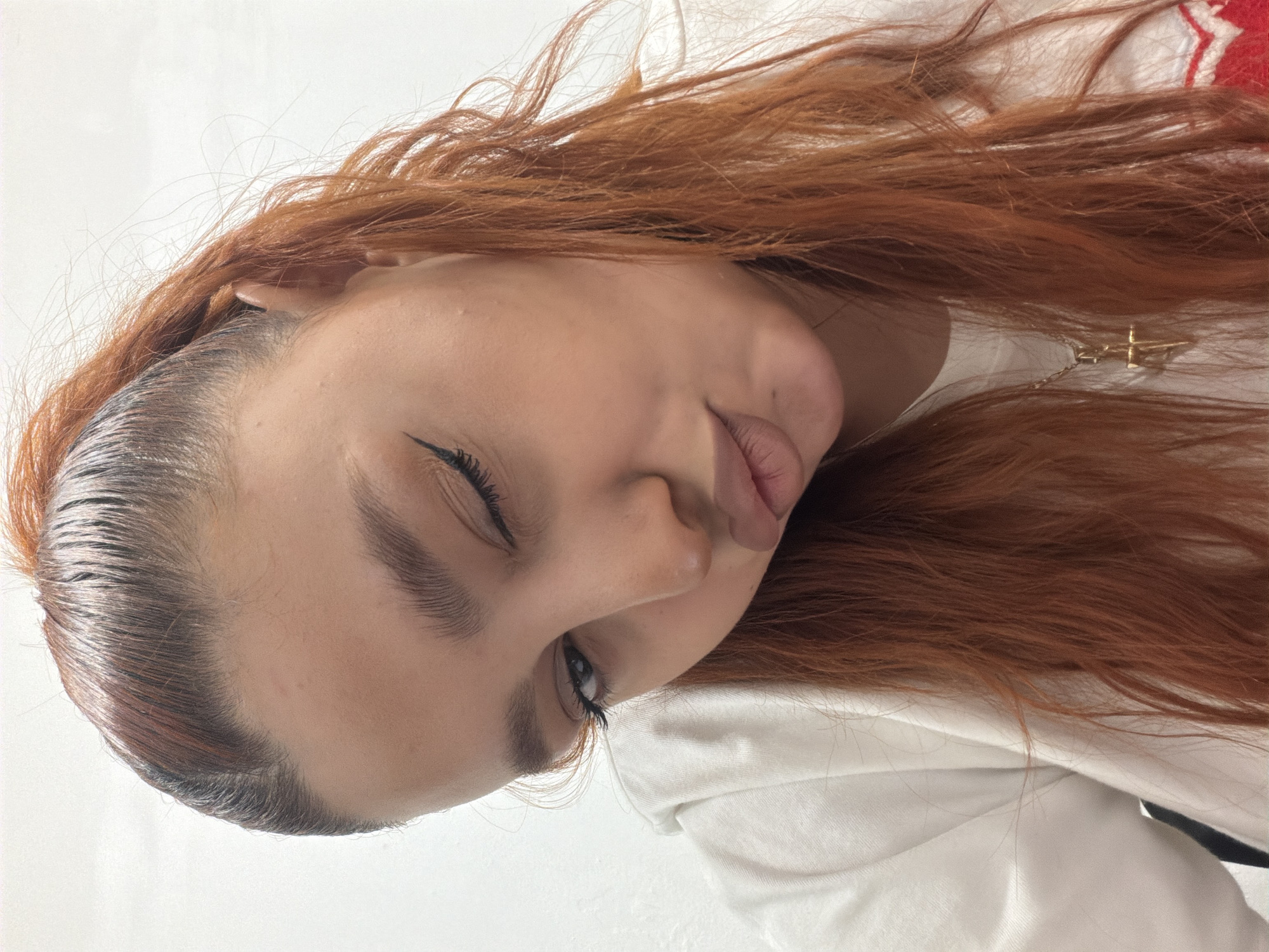 A close-up, angled portrait shows a person with fair skin, closed eyes, and auburn hair. The hair is swept back at the forehead, revealing an auburn hairline. A black eyeliner is visible on the closed eye. The person is wearing a white hoodie, and a gold necklace with a cross pendant is partially visible. The background is plain white. There is no evidence to suggest this photograph was taken in Yerevan, Armenia.