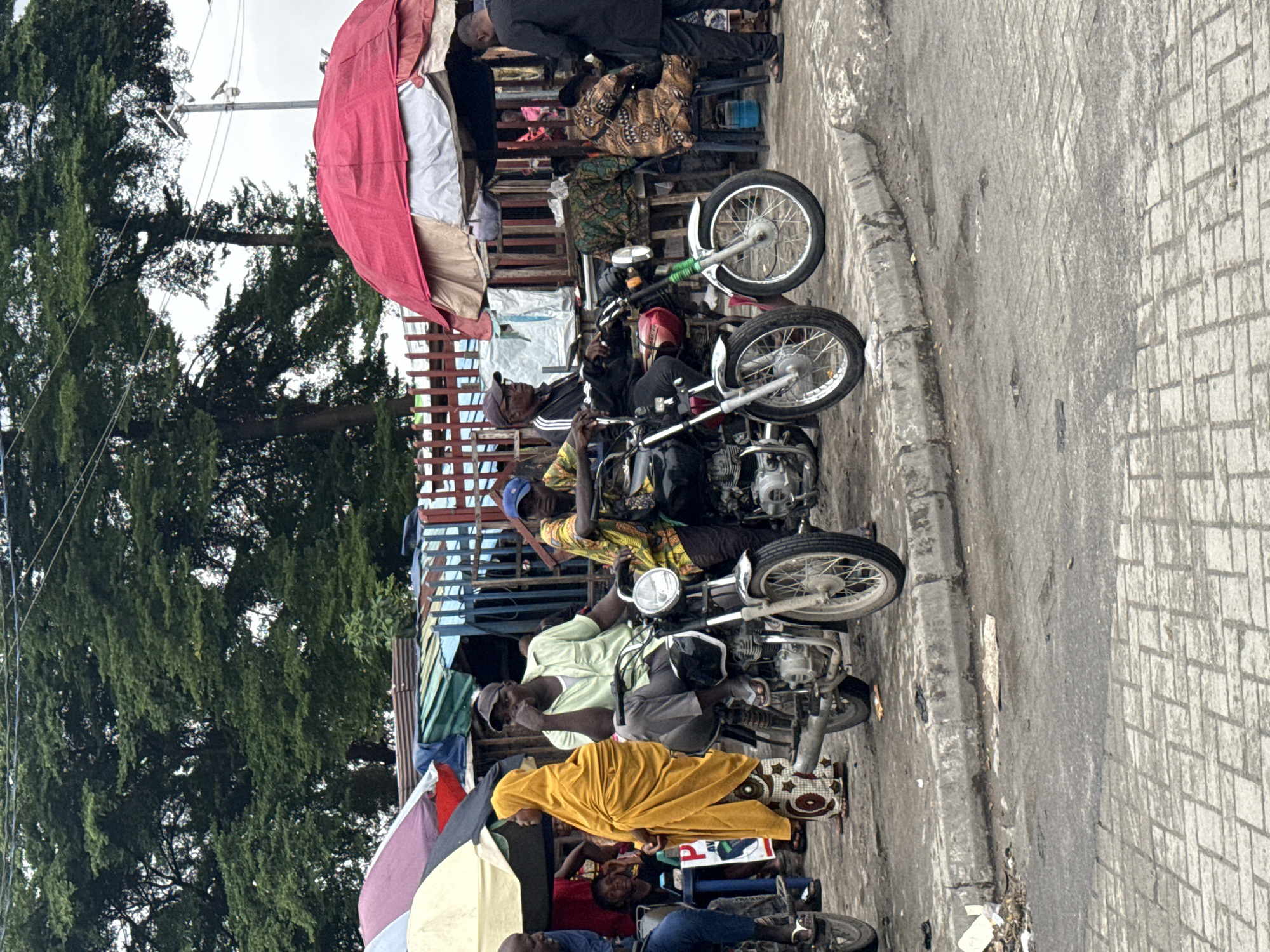 The image depicts a street scene in Ado, Nigeria, with several individuals and motorcycles. In the foreground, a man in a light green shirt and grey pants sits on a motorcycle, resting his chin on his hand. Next to him, another man in a colorful patterned shirt and blue cap sits on a similar motorcycle. To the left, a woman in a bright yellow hijab stands, and a person in blue jeans is partially visible on a motorcycle. In the background, there are market stalls with various awnings, including a large red one, and abundant trees. Several other people are seen milling around the stalls and motorcycles, some seated and others standing. The ground is paved with interlocking bricks and asphalt, with some debris visible. The overall atmosphere suggests a busy, local marketplace or transportation hub.
