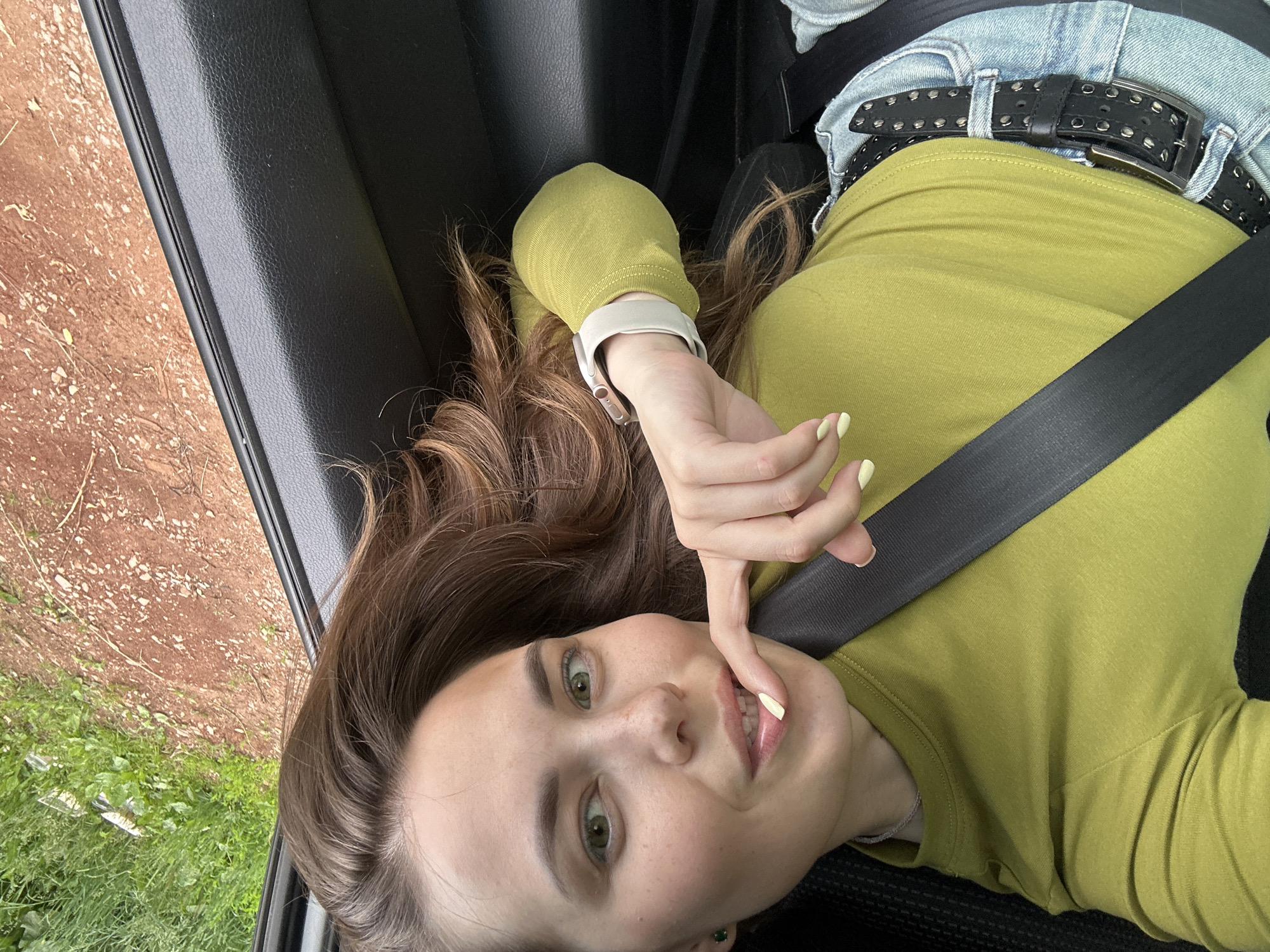 The image shows a young woman with brown hair and green eyes, wearing a lime green long-sleeved shirt, blue jeans, and a black studded belt. She is sitting in a car, with a seatbelt across her chest, and resting her head against the window. Her finger is touching her lips, and she is looking at the camera. The car door is open, revealing a view of a rural area with green grass and a dirt road. The scene appears to be set in Ilyinsky, Russia, as indicated by the available context.