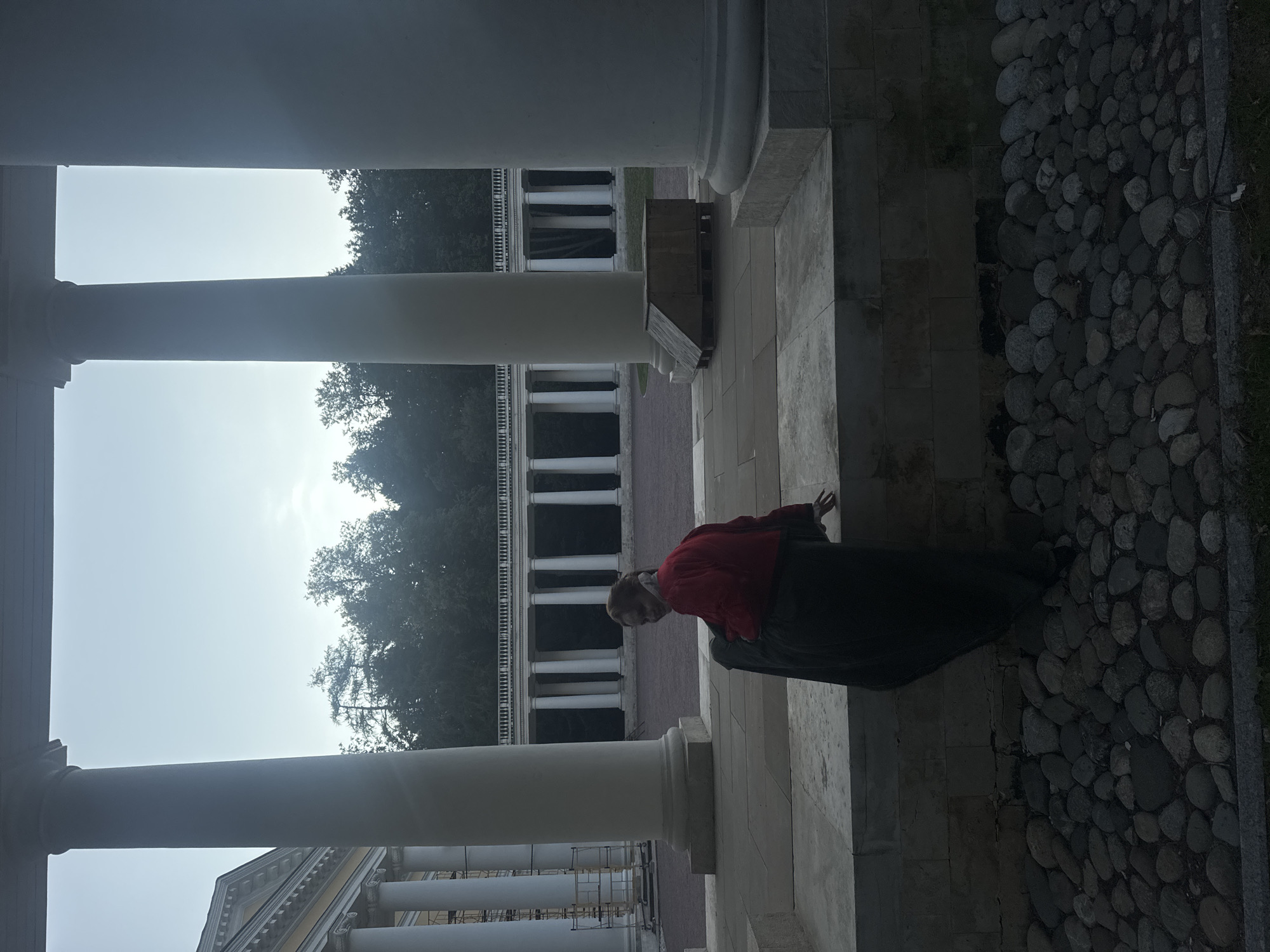 In the city of Arkhangel'skoe, Russia, a young woman is seated on a stone ledge, her back partially turned towards the camera. She is wearing a red top and a long dark skirt. The backdrop is an architectural ensemble featuring a colonnade of large, white pillars framing a distant building and a line of trees under a pale sky. In the foreground, the ledge is bordered by a collection of smooth, grey stones. The overall scene suggests a historical or classical setting.