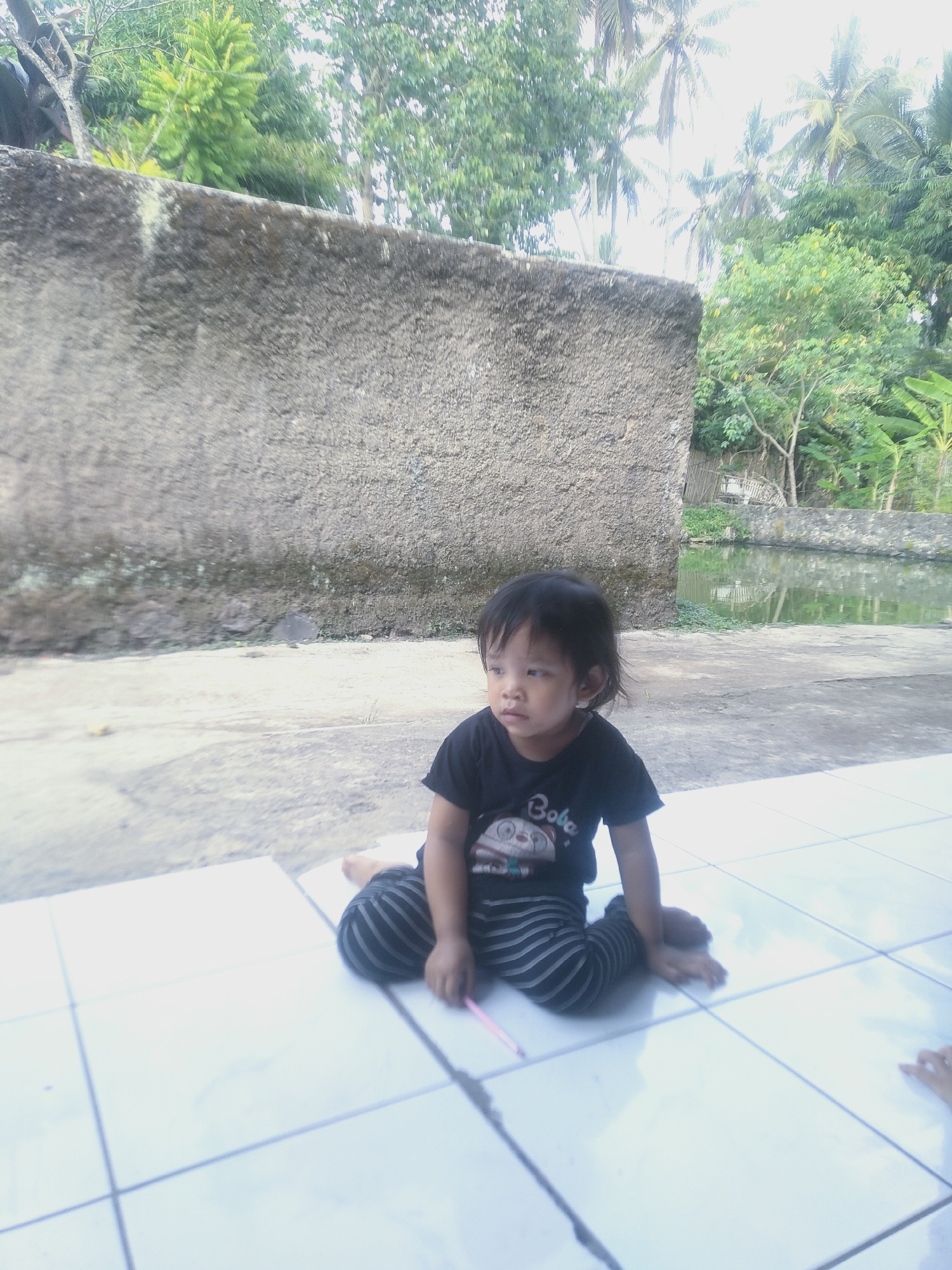 A young child with dark hair sits cross-legged on a white tiled floor in front of a rough, grey concrete wall. The child wears a black t-shirt with a cartoon image and text, and striped pants. The background shows lush green foliage, palm trees, and a body of water. The scene appears to be outdoors, possibly in a park or residential area in Bojong, Indonesia.