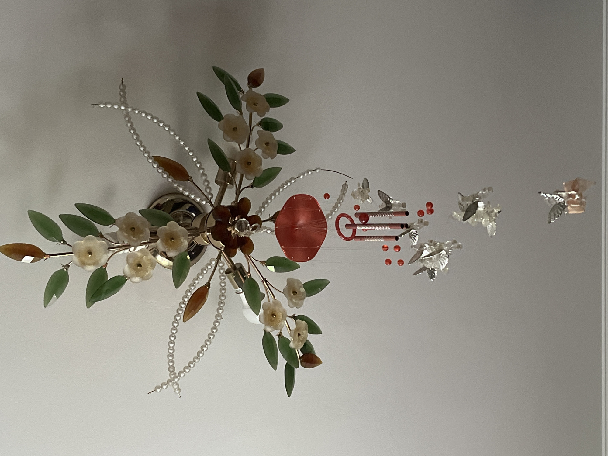 The image displays a decorative ceiling fixture, possibly a light or a mobile, suspended against a plain, light-colored ceiling. The main body of the fixture features gold-toned metallic branches adorned with numerous light-colored, translucent, carved floral elements and green, translucent, carved leaf elements. Strands of small, white, spherical pearls arc outwards from the central fixture. Several faceted, amber-colored elements are also integrated into the design.
Hanging from the central fixture is a red, translucent, round, umbrella-shaped ornament. Below this, two red-capped cylindrical metallic chimes are suspended. A cluster of small, round, red beads is visible near the chimes. Further below, several delicate, clear glass or crystal butterflies with silver-toned metallic bodies and wings are suspended at varying lengths, creating a cascading effect. No people or specific identifying environmental features are present to determine a location or city such as Minsk, Belarus.