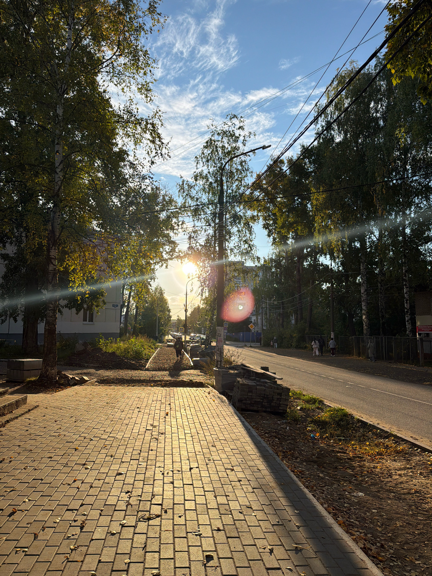 The image depicts an autumnal scene on a paved sidewalk in Chaikovsky, Russia, bathed in the warm, low-angled light of the sun. The sidewalk, constructed of rectangular pavers, is partially covered with fallen leaves. To the left of the sidewalk is a grassy area and a building, partially obscured by trees. The foreground shows a stack of paving stones, suggesting ongoing construction or maintenance. Further down the sidewalk, a lone figure walks away from the camera, silhouetted against the bright sunlight.
Beyond the sidewalk is a paved road with several cars and a few more figures walking on the opposite side. Trees line the road, their leaves displaying autumnal colors. Overhead, utility lines are visible against a blue sky with scattered, wispy clouds. A lamppost stands near the center of the image. The sun is bright and positioned high in the sky, creating lens flare and a reddish circular artifact. The overall impression is of a quiet, late afternoon in a residential area during the autumn season.