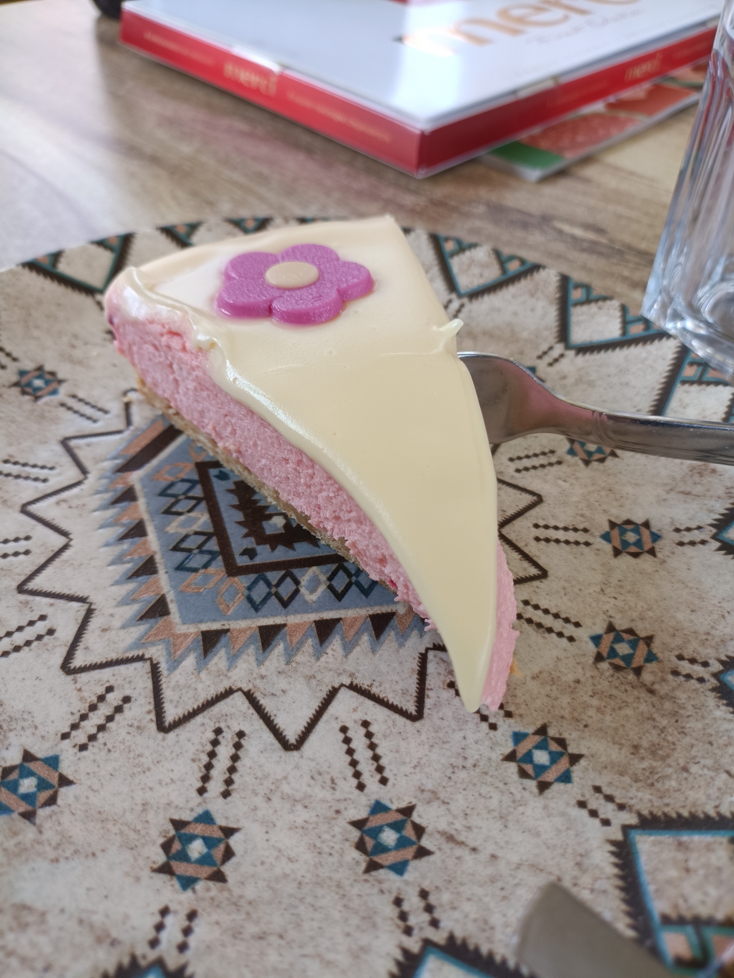 A slice of pink cheesecake with a white icing layer and a purple flower decoration sits on a patterned plate. A fork pierces the cake. A glass and a book with the word "merci" are visible in the background. The scene is on a table, likely indoors. The location in the city of Plovdiv, Bulgaria is not determined from the image.