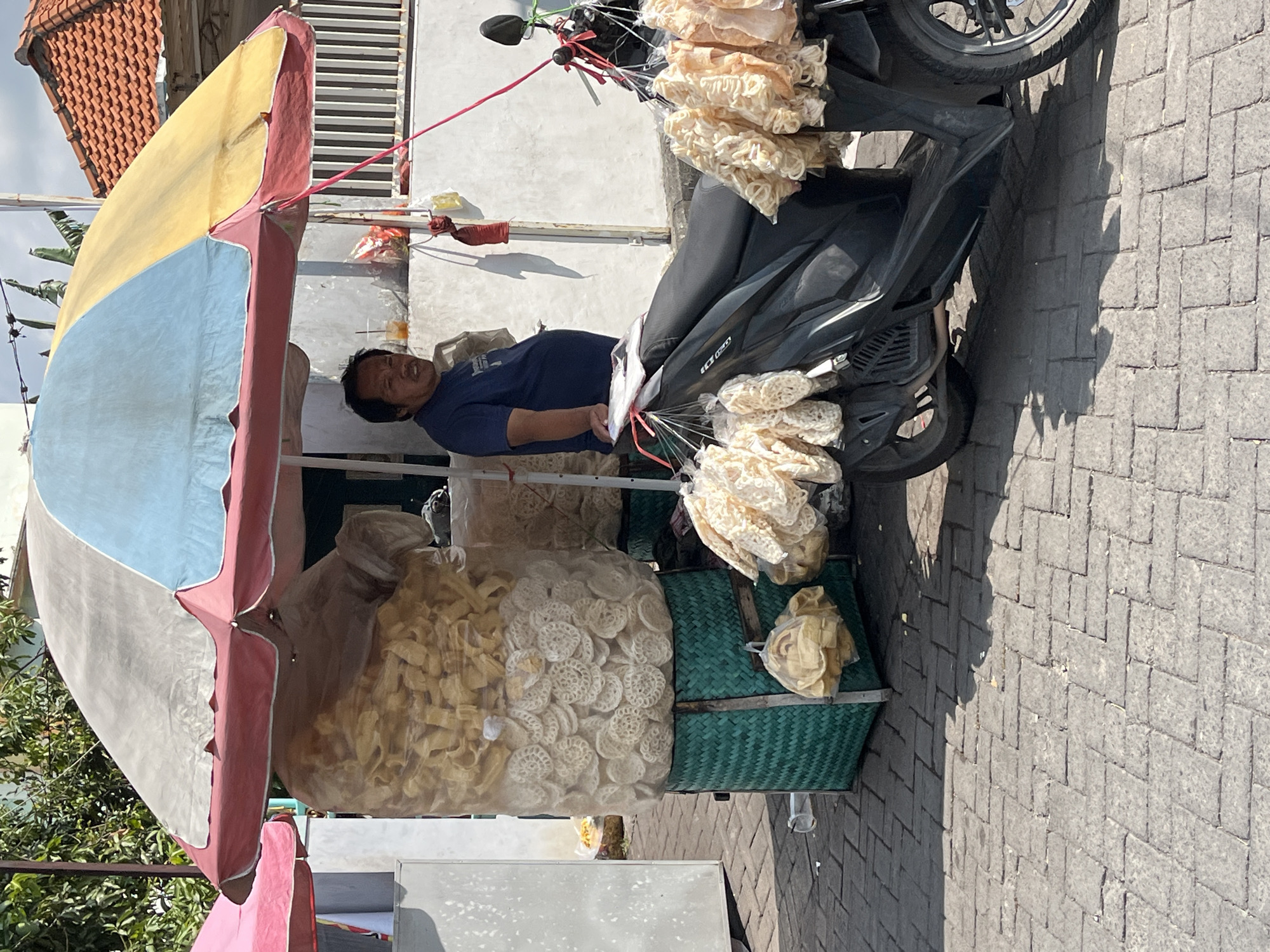A street vendor stands behind a stall selling large quantities of snacks, likely crackers or chips, displayed in clear plastic bags on a sunny day in Kalisampurno, Indonesia. The stall is covered by a large, multicolored umbrella, predominantly yellow and light blue, with red trim. A green woven basket supports a tall, clear plastic bag filled with light-colored, circular snacks. To the right, a black motorcycle is parked, also adorned with bunches of these snacks hanging from its handlebars and side. The vendor, a man with dark hair and wearing a dark blue t-shirt, is positioned behind the stall, looking towards the viewer. The ground is paved with interlocking gray bricks. In the background, a white wall and a building with a tiled roof are visible, suggesting an urban or semi-urban setting. The scene is illuminated by bright sunlight, casting distinct shadows on the ground.