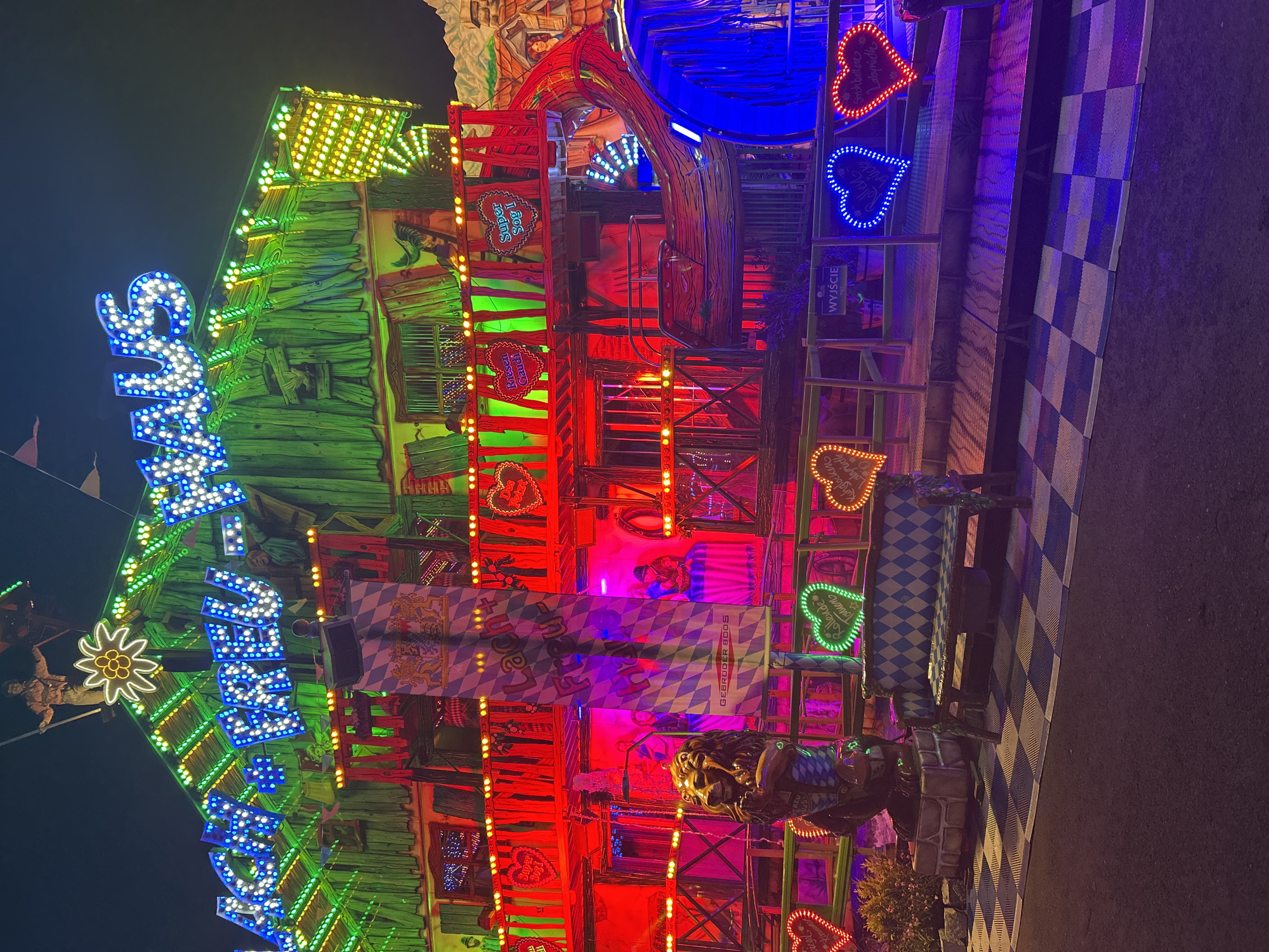 This is a nighttime, medium shot of a brightly lit amusement park attraction in Szczecin, Poland. The attraction, a large wooden structure adorned with numerous colorful lights, is illuminated with neon blues, greens, and reds. A large sign with blue neon letters reads "LACH+FREU+HAUS". The structure features multiple levels with railings, and decorative elements such as heart-shaped lights and checkered patterns are visible. In the foreground, a bench with a checkered design sits on a platform. A lion statue stands to the left of the bench. The ground is a checkered pattern of black and white tiles. The background is dark, indicating nighttime.