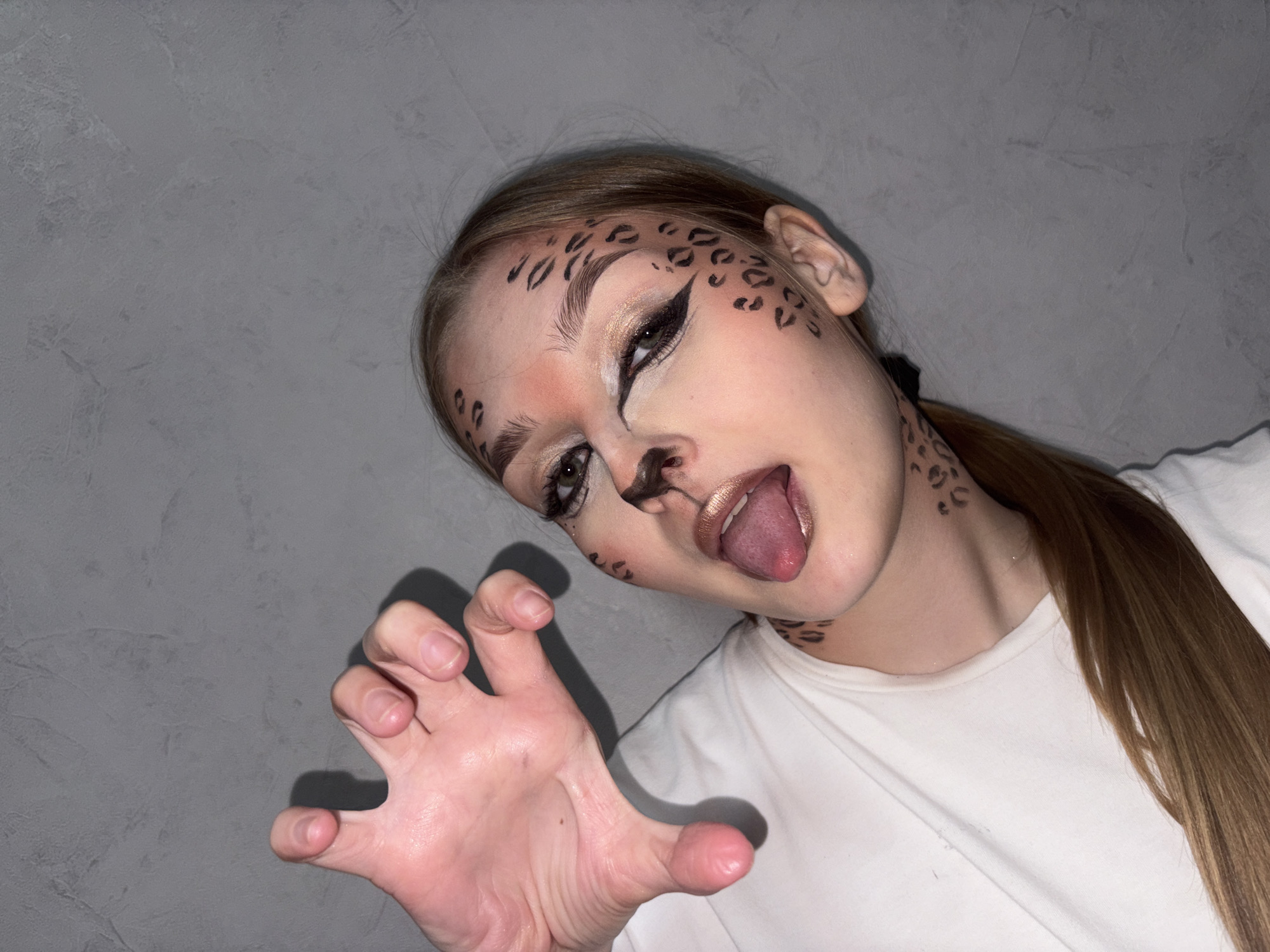 A young woman with light skin is posed indoors against a light gray, textured background. She is wearing a white t-shirt. Her makeup is designed to resemble the markings of a leopard or cheetah, with black spots painted around her eyes, forehead, cheeks, and neck. Her eyes are heavily lined with black eyeliner, and she has gold eyeshadow. Her lips are a metallic gold color. She is sticking out her tongue and has her hand outstretched in a claw-like gesture. Her long, light brown hair is pulled back in a ponytail. The location is Chaikovsky, Russia.