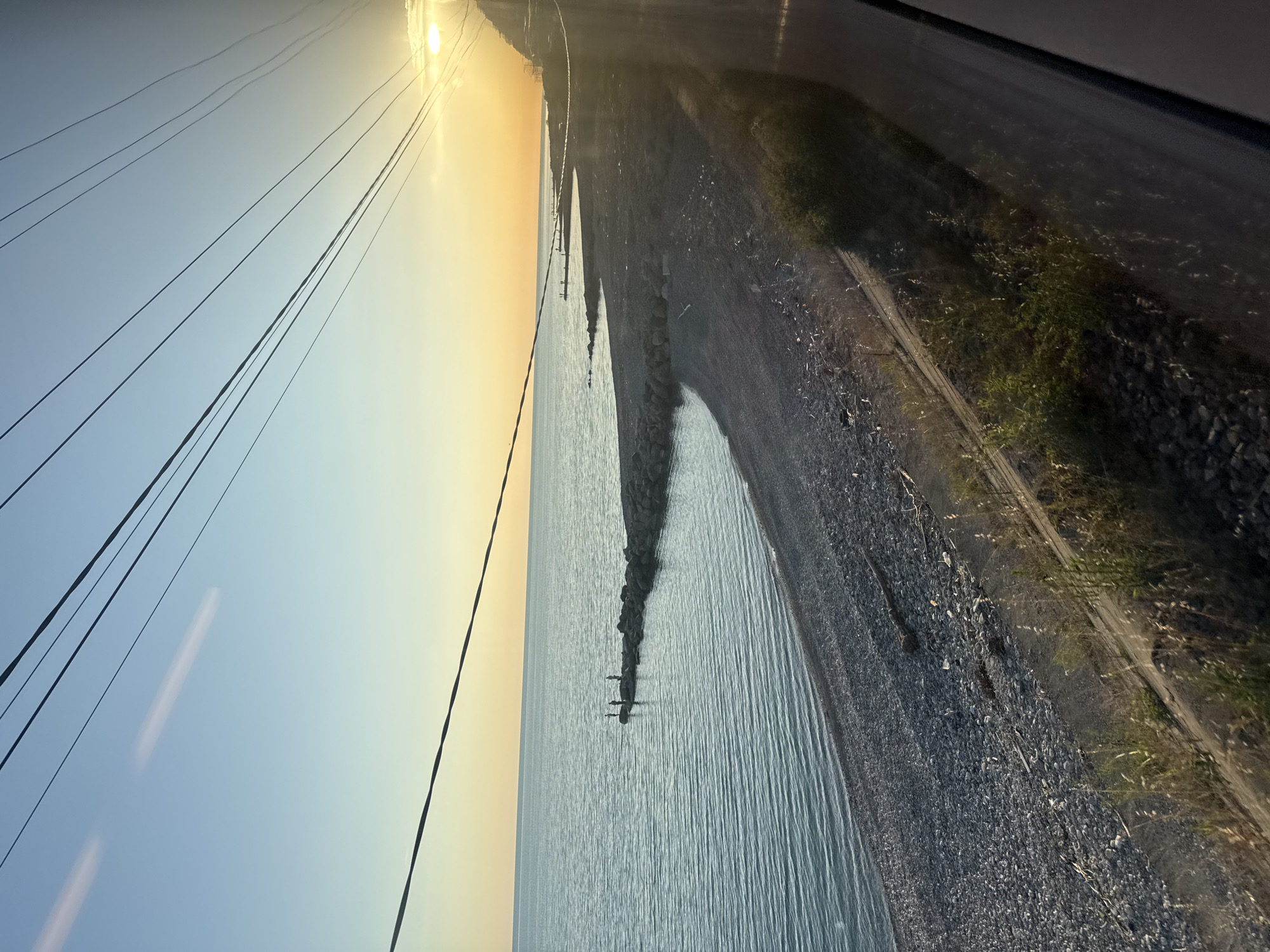 This landscape image captures a serene coastal scene at either sunrise or sunset, characterized by a vibrant sky and calm waters. The sun is a bright, intense orb located in the upper right quadrant, illuminating the sky with warm yellow and orange hues that fade into a soft blue towards the left.
Multiple dark power lines crisscross the sky diagonally from the lower left to the upper right. Below the horizon, a placid body of water, likely the Black Sea given the Sochi, Russia context, stretches across the middle ground, reflecting the sky's gentle light and showing subtle ripples on its surface.
Along the shoreline, a dark, pebbly or rocky beach separates the water from the land. Several dark, elongated structures, appearing to be groynes or breakwaters, extend into the sea from the shore. To the right of the beach, a dark road or path runs parallel to the coast, with patches of sparse green vegetation, including grass and small bushes, growing alongside it.
The scene is devoid of people or visible activity, contributing to a peaceful and natural ambiance. The weather appears clear with no clouds. There is no visible text in the image.