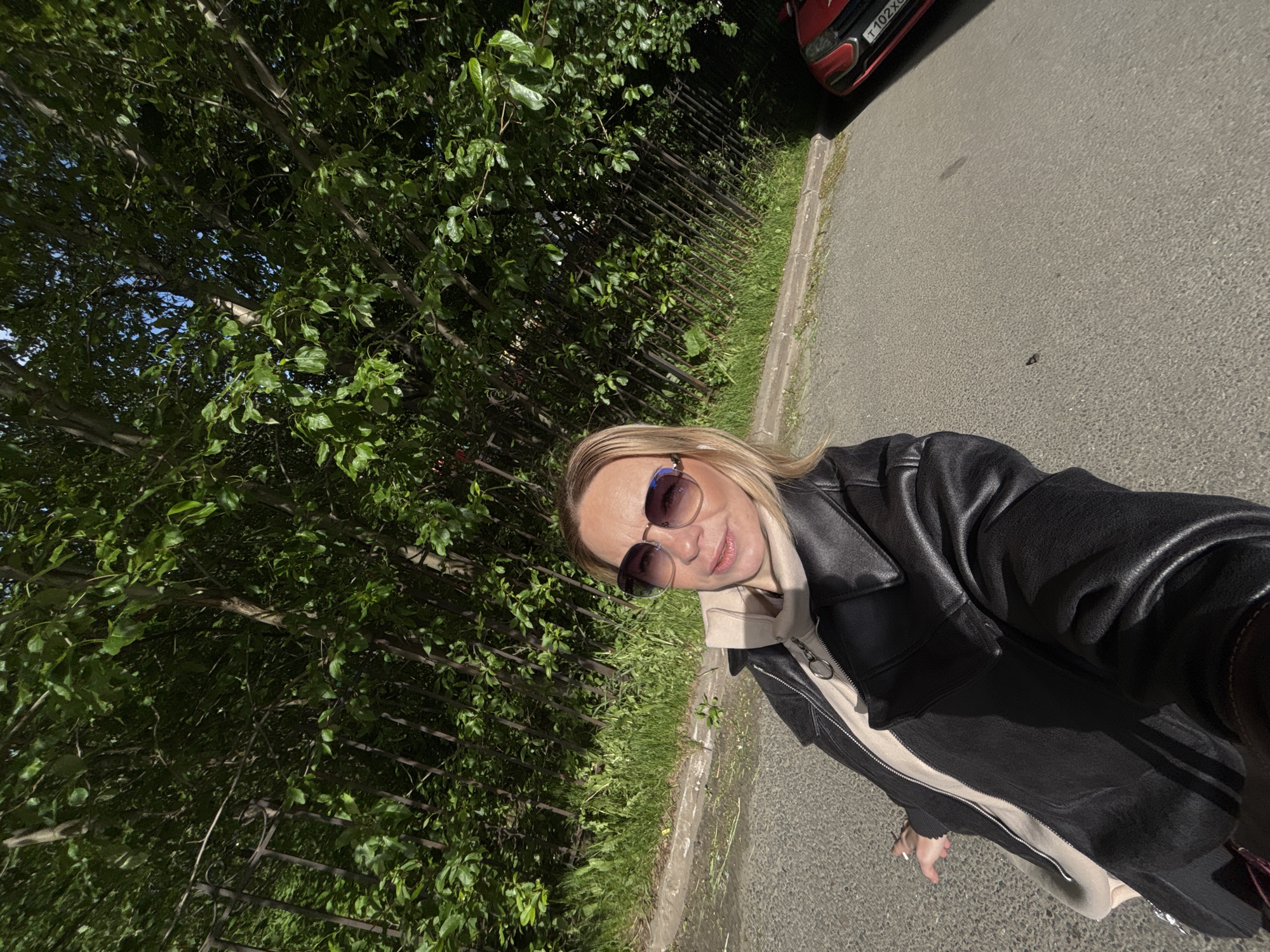 A woman wearing sunglasses and a black leather jacket is standing on a paved surface, possibly a sidewalk or road, next to a tree and a fence. A red car is partially visible in the upper right corner. The license plate of the car reads "T 102 XO," which suggests the location is Russia, possibly Saint Petersburg. The surroundings include greenery suggesting a park or tree-lined street. The woman appears to be taking a selfie.