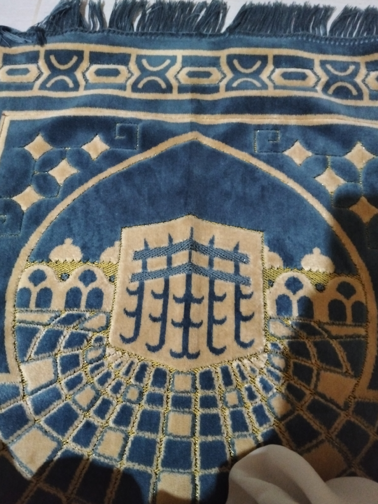 A close-up, overhead view of a blue and tan prayer rug with intricate patterns. The rug features a central design resembling the Kaaba in Mecca, surrounded by arched doorways and a radiating checkerboard pattern. The rug has a fringed edge at the top. The lighting suggests an indoor setting, with shadows cast on the rug. The specific location in Payagaboh, Indonesia, is not visible in the image itself, but the prayer rug indicates a likely Islamic context.