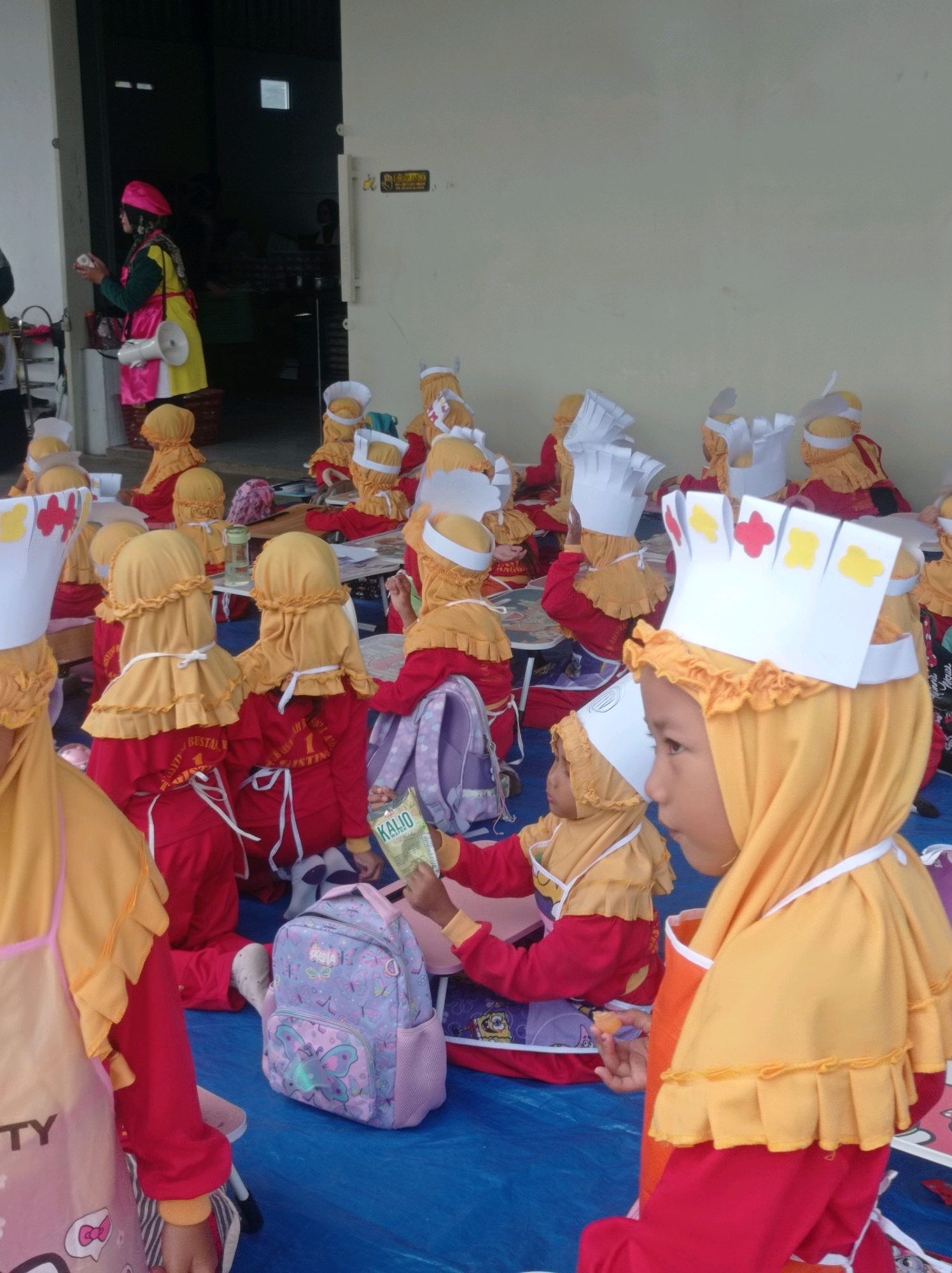 In Gisting Bawah, Indonesia, a group of young children, likely participating in a cooking or creative activity, are gathered outdoors on a blue tarp. Most of the children are wearing matching red shirts, yellow headscarves, and paper chef hats, some adorned with colorful paper shapes. Several children are holding or interacting with objects, including a purple backpack with butterfly designs, a box of "KALIO" brand crackers, and what appears to be food items. A woman in a colorful yellow, pink, and green outfit, possibly an instructor or supervisor, stands near a doorway in the background, holding a white megaphone. The environment suggests a community or school event, with a building wall and a doorway visible.