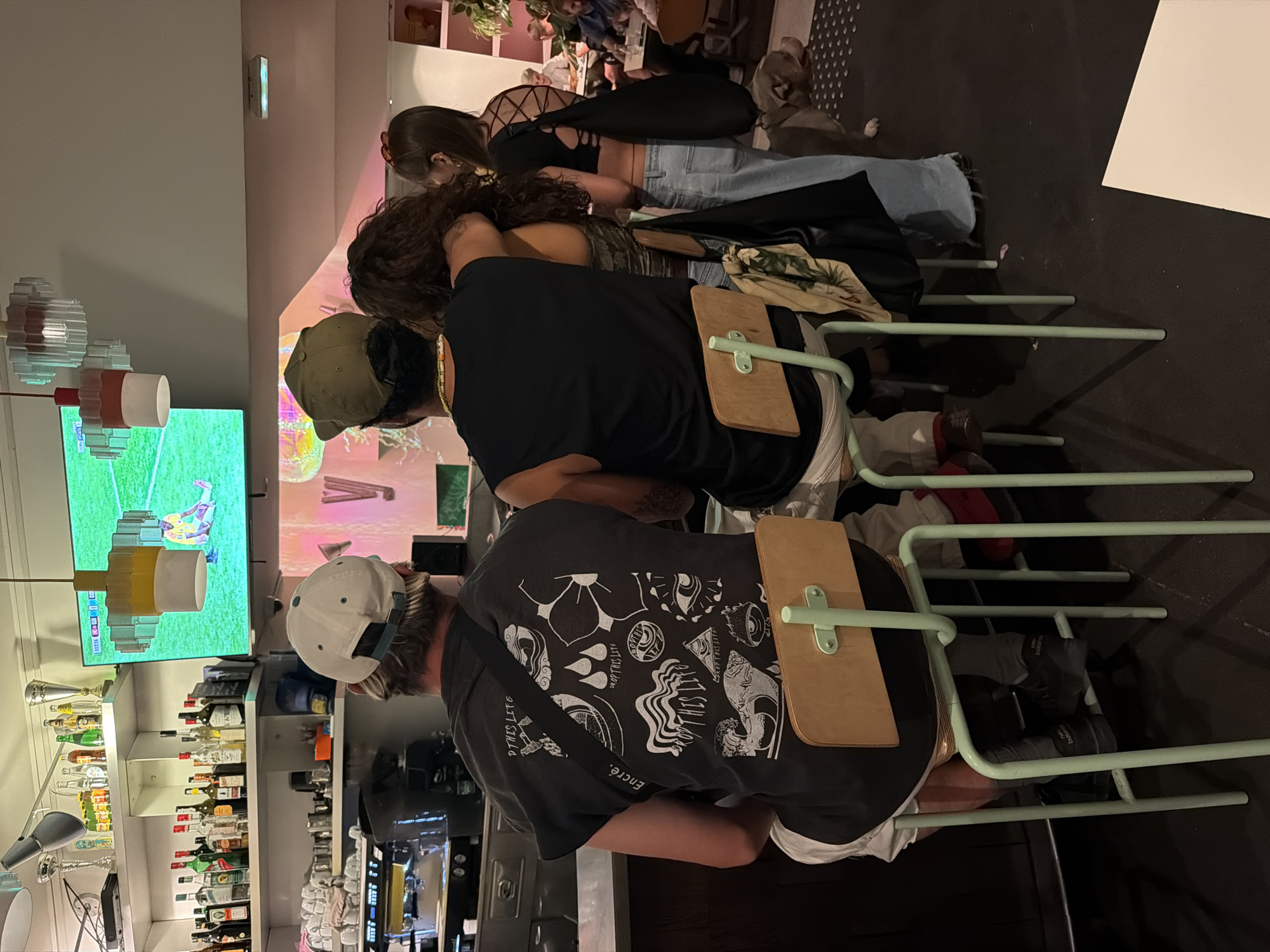 In what appears to be a dimly lit bar or restaurant in Lyon, France, three individuals are seated at a counter, facing away from the camera. The person in the foreground, wearing a dark t-shirt with a graphic print and a white baseball cap, has their back to the viewer. To their right, another individual in a black tank top and a green baseball cap is seated, with their arm around the person in the middle, who is wearing a camouflage-patterned top. Further to the right, a fourth person, a woman with dark hair, is also seated. The background features shelves stocked with bottles, a television displaying a soccer game, and a barista machine. A dog stands on the floor to the right of the seated individuals. The overall ambiance suggests a casual gathering or a night out.