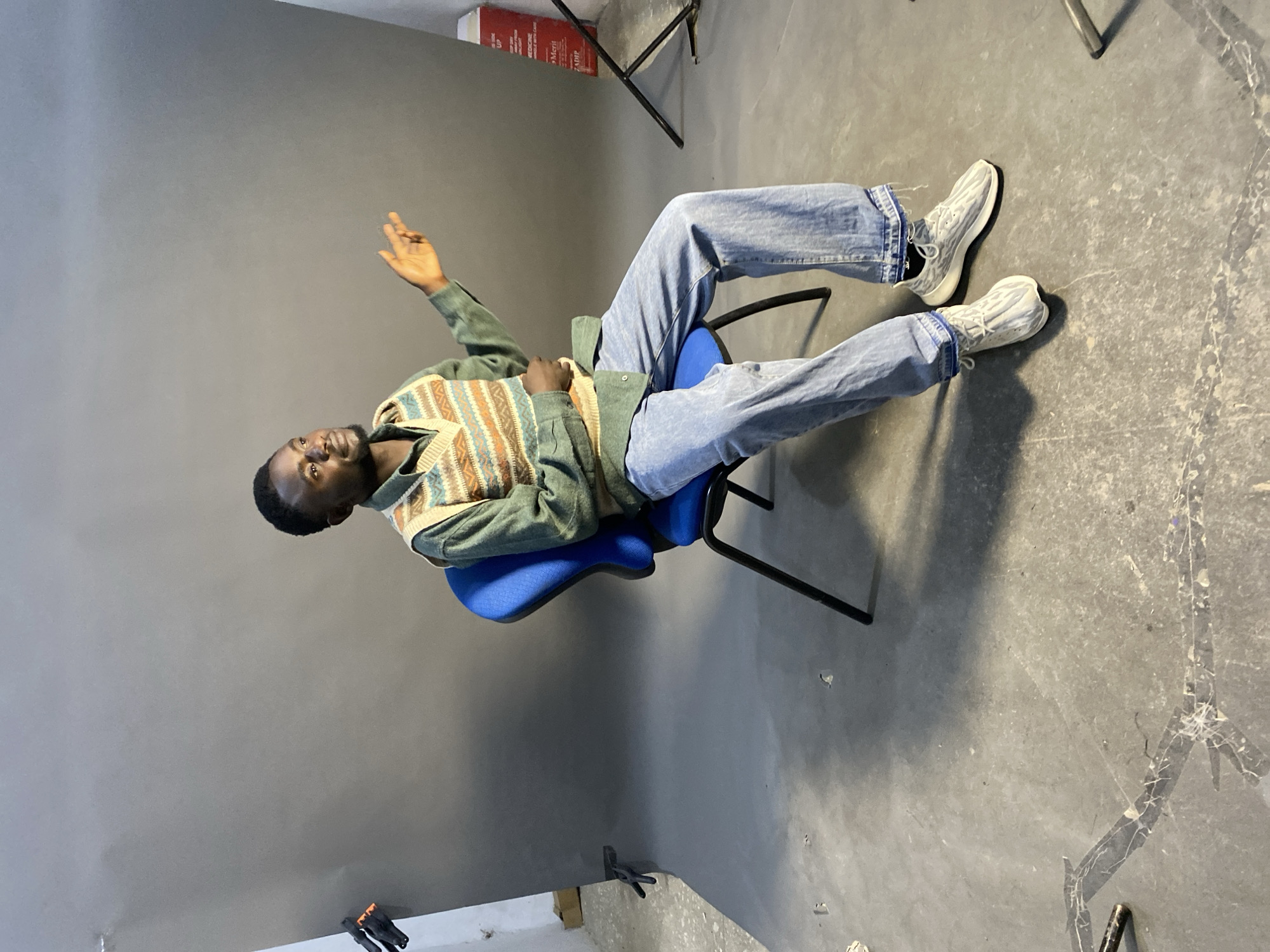 A young Black man is sitting in a blue office chair in what appears to be a photography studio. He is leaning back, facing the camera, with his right arm raised. He is wearing light blue jeans, white sneakers, a green shirt, and a patterned sweater vest. The studio has a gray backdrop and a concrete floor. There is photography equipment visible, including light stands. The location in Jos, Nigeria is assumed, but not visually confirmed.