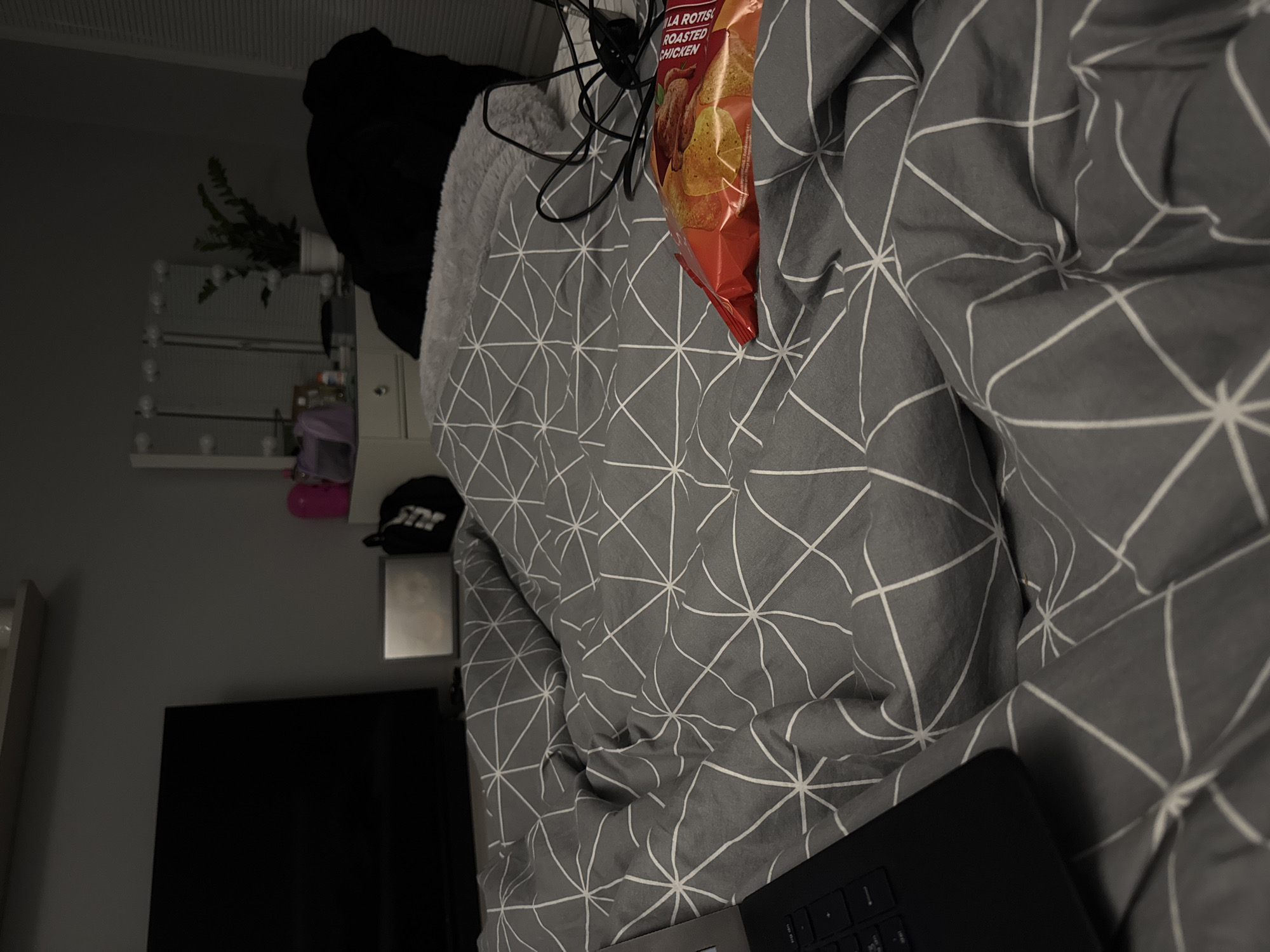 The image depicts a disheveled bed covered with a grey and white geometric-patterned duvet. A bag of "Lay's Rotisserie Roasted Chicken" flavored potato chips is resting on the bedspread. A black laptop sits partially open on the duvet. In the background, there is a black hoodie, a white dresser with a light-up mirror, a black baseball cap, and a television. The room is poorly lit. There are no identifiable elements that would place this image in Sofia, Bulgaria.
