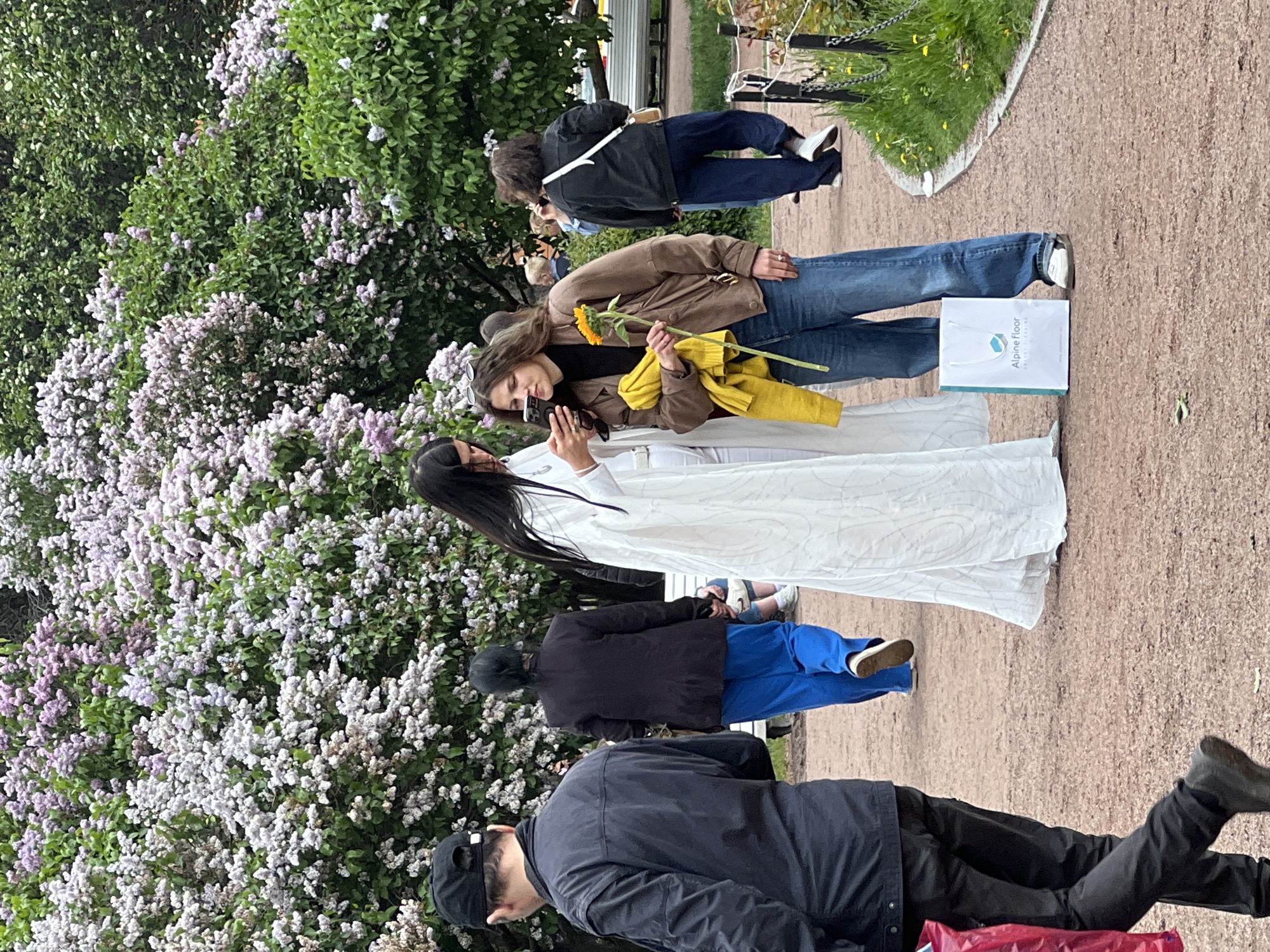 The image shows a scene in the city of Saint Petersburg, Russia. In the background is a lush lilac bush with blooming purple and white flowers. A woman in a long white dress and another woman holding a sunflower are posing for a photo. The photo is being taken by a third woman who is holding a smartphone. To the right of the posing women is a shopping bag. The foreground shows pedestrians, likely also tourists. The ground is covered in a reddish-brown gravel or compressed stone.