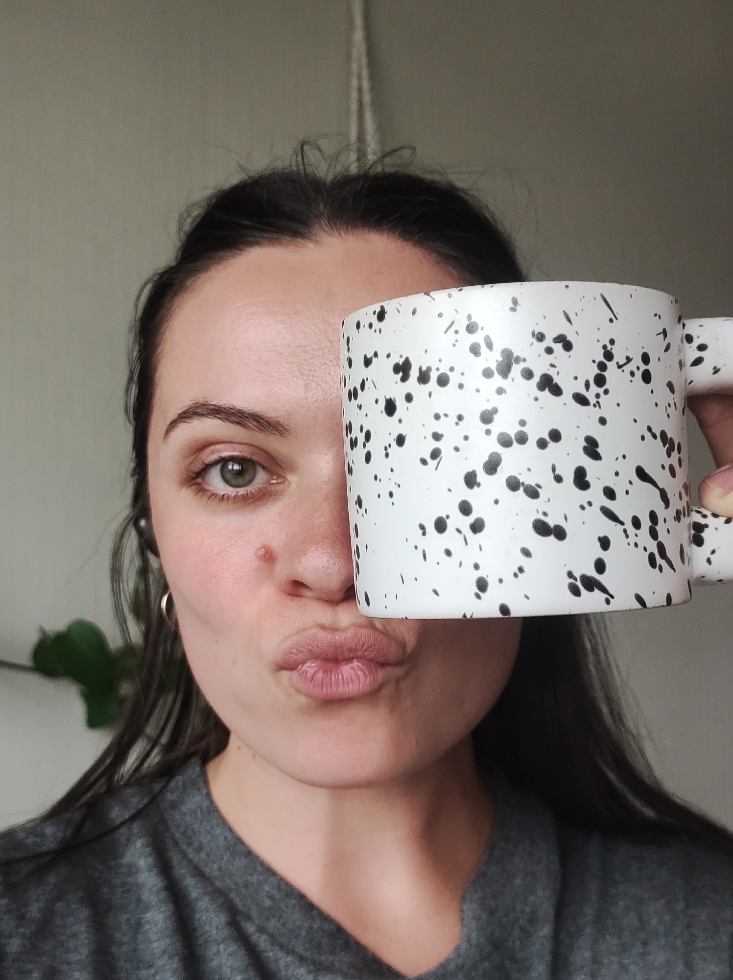 A woman, partially obscured by a white speckled mug held to her face, puckers her lips towards the camera. Only her right eye, eyebrow, cheek, nose, and mouth are visible. She has dark hair pulled back and is wearing a grey t-shirt. The background is a plain, light-colored wall. There is no indication of Chaikovsky, Russia in this image.