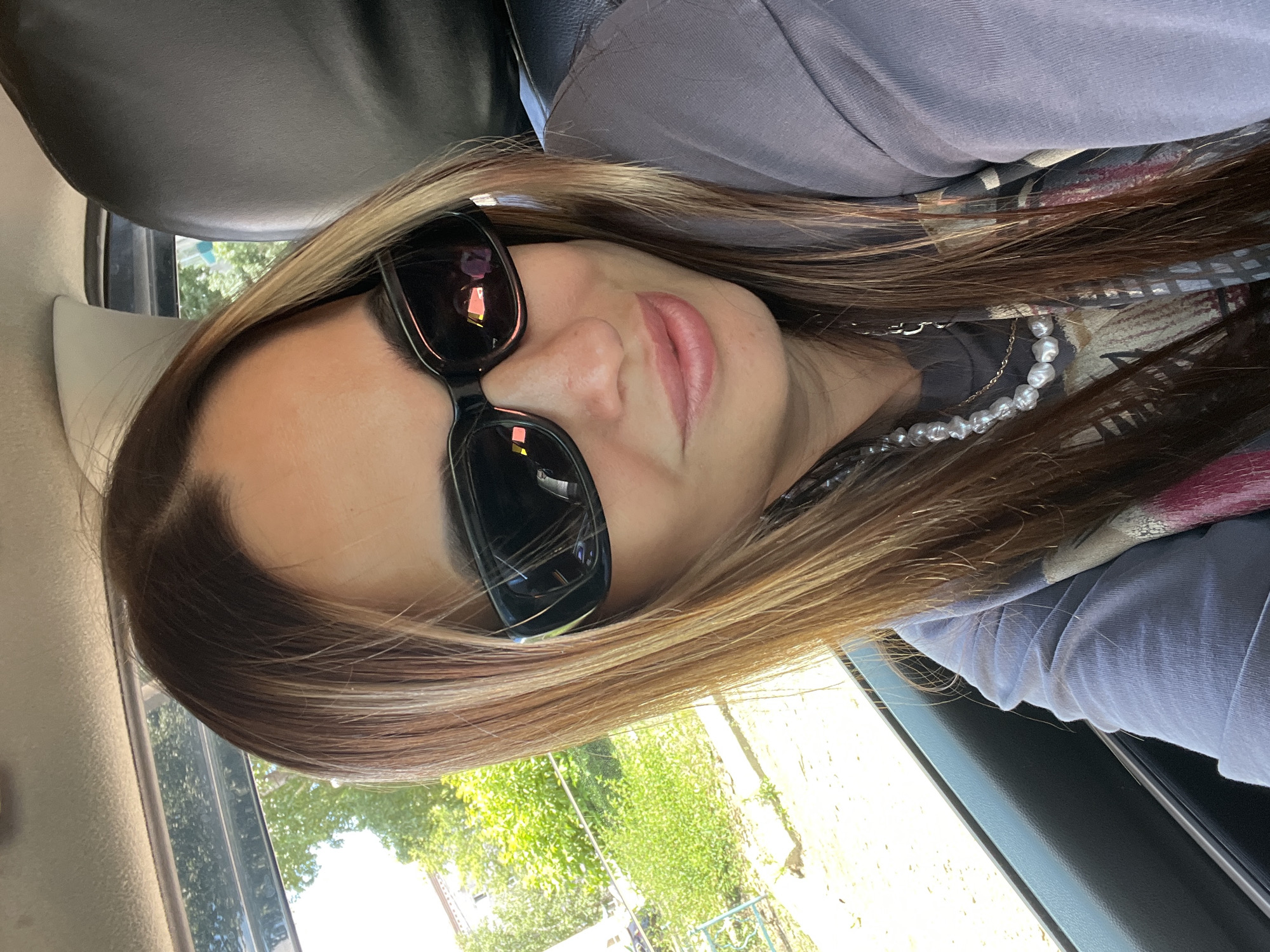 A woman wearing sunglasses and a pearl necklace is sitting in a car. She has long, highlighted brown hair and is wearing a gray t-shirt. The car appears to be parked outdoors, with lush green trees and bushes visible through the window. The scene is lit by natural daylight, suggesting it is daytime.