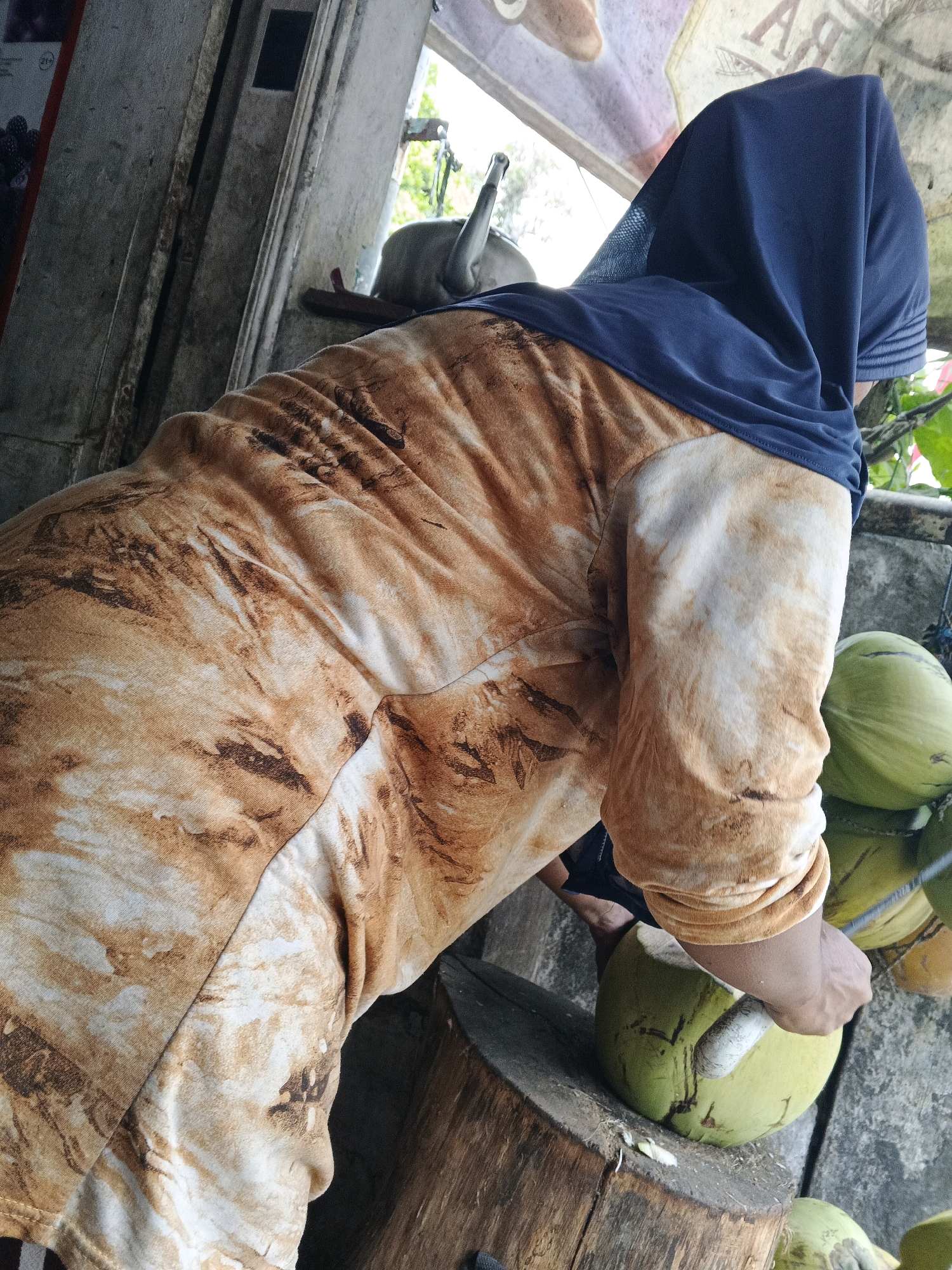 The image depicts a street vendor in Sukabumi, Indonesia, preparing coconuts. The vendor, wearing a blue headscarf and an orange and brown patterned shirt, is in the process of husking a green coconut with a knife on a wooden chopping block. Several other coconuts are visible in the background, some of which are stacked and appear ready for sale. The scene suggests a typical roadside stall, with a rough wooden structure to the left and an open doorway or window providing a glimpse of the outdoors. The lighting indicates it is daytime.