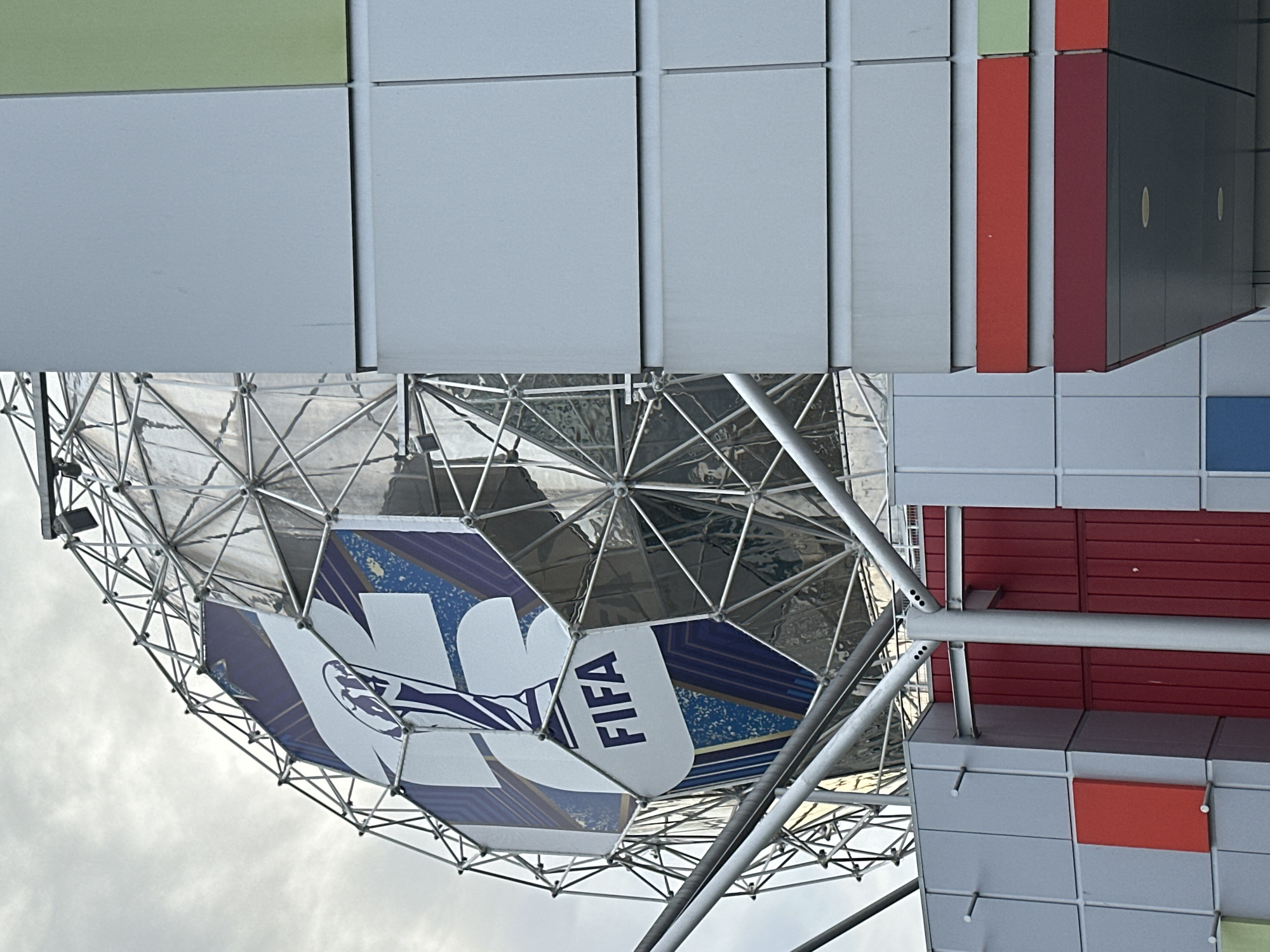 The image shows a geodesic sphere with a FIFA World Cup promotional design. The design features the FIFA logo and branding elements. The sphere is supported by a network of metal beams. The structure is located near a building with a modular panel facade featuring a variety of colors, including gray, green, red, and blue. The image was taken in Vancouver, Canada.