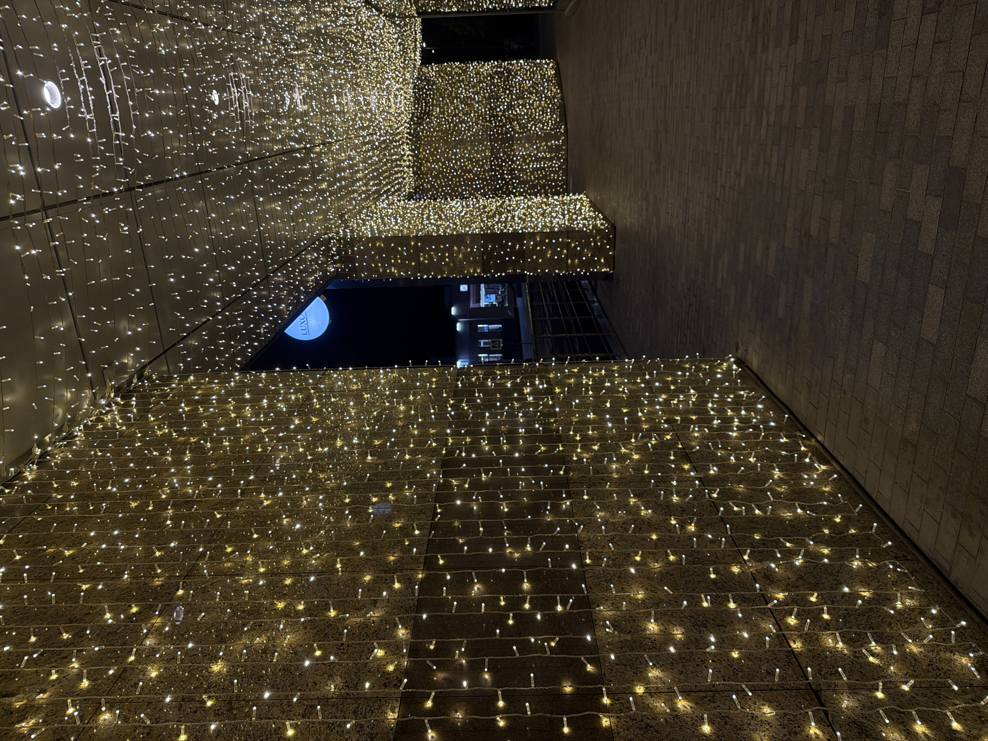The image shows an exterior view of a building or structure adorned with numerous small, warm-toned fairy lights, creating a dense and illuminated display. The lights are attached to a surface that appears to be made of dark brick or stone. The composition is taken from a low angle, looking upwards towards a gap or opening in the structure, revealing a dark sky with a partial view of another building in the background. The scene is likely taken at night, given the darkness and the reliance on artificial lighting. The building on the right appears to be a light brown brick and doesn't have any lights. There is nothing to confirm the location.