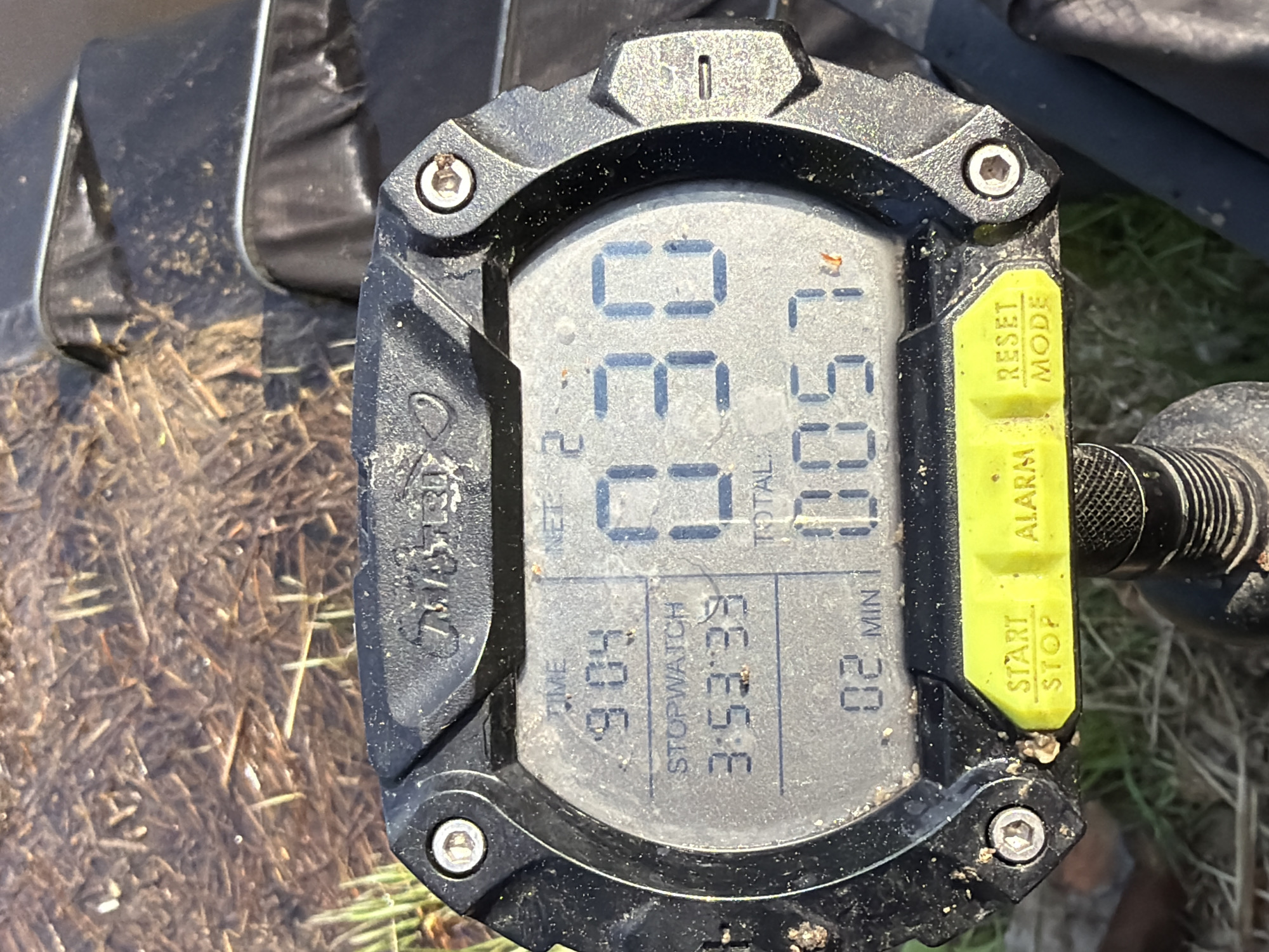 The image depicts a close-up view of a digital watch lying on a surface covered with straw and dirt, possibly outdoors. The watch, a Tekstiel model, has a black, rugged casing with a yellow control panel on the right side, featuring buttons labeled "RESET MODE" and "START STOP ALARM". The watch display shows the following data: Time: 9:04, Stopwatch: 3:53:33, Net: 663, Total: 0057, and 02 Min. The watch appears dusty and has debris stuck to it. The location in Chaykovsky, Russia, is not discernible from the image alone.