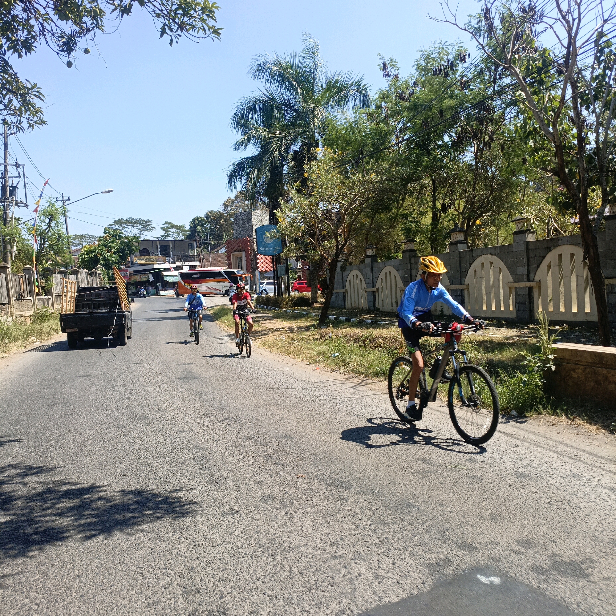 The image depicts a sunny day scene on a paved road, likely in Kebondowo, Indonesia, with a line of three cyclists. The cyclist in the foreground, an adult male wearing a blue long-sleeved shirt, black shorts, and a yellow helmet, is riding a black bicycle with a red water bottle cage. To his left and further down the road, two other cyclists are visible: one in a red shirt and black shorts, and another in a blue shirt and black shorts, both wearing helmets. A black pickup truck with a wooden frame is parked on the left side of the road. In the background, there are buildings, trees, and a large bus. The environment includes a clear blue sky, trees with green foliage, and some dry vegetation along the roadside. The road appears to be in moderate condition, with some cracks and unevenness. The overall impression is of an outdoor recreational activity taking place in a semi-urban or rural setting.