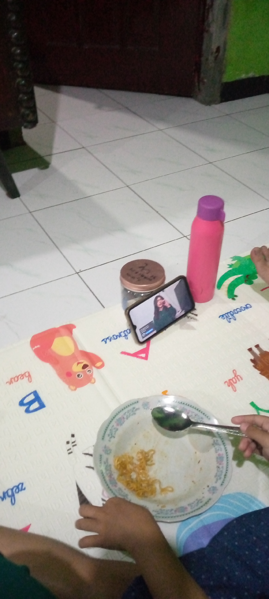 A close-up shot captures a meal on a play mat on the floor, likely in a residential setting in Sumberwono, Indonesia. A partially eaten plate of noodles sits in the foreground, with a spoon held by a person's hand poised to take another bite. To the left of the plate, a red cartoon bear is printed on the mat, with the word "bear" written below it. To the right, a pink water bottle with a purple lid stands upright next to a glass jar with a brown lid. A smartphone is propped up, displaying a woman on a video call. The play mat also features printed words like "crocodile" and "zebra". The background shows a tiled floor and the corner of a dark wooden cabinet. The scene suggests a casual mealtime, possibly for a child, with the woman on the phone perhaps present virtually.