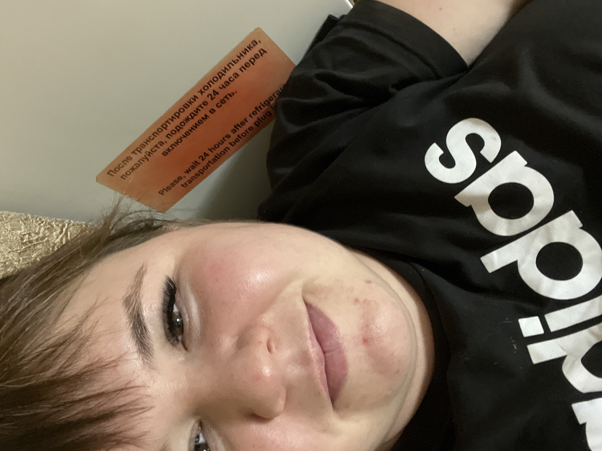 A person lies on a surface, with an orange label attached to a refrigerator cautioning in both Russian and English to wait 24 hours after transportation before plugging in. The person is wearing a black Adidas shirt. The image does not provide details about the location in Chaikovsky, Russia.