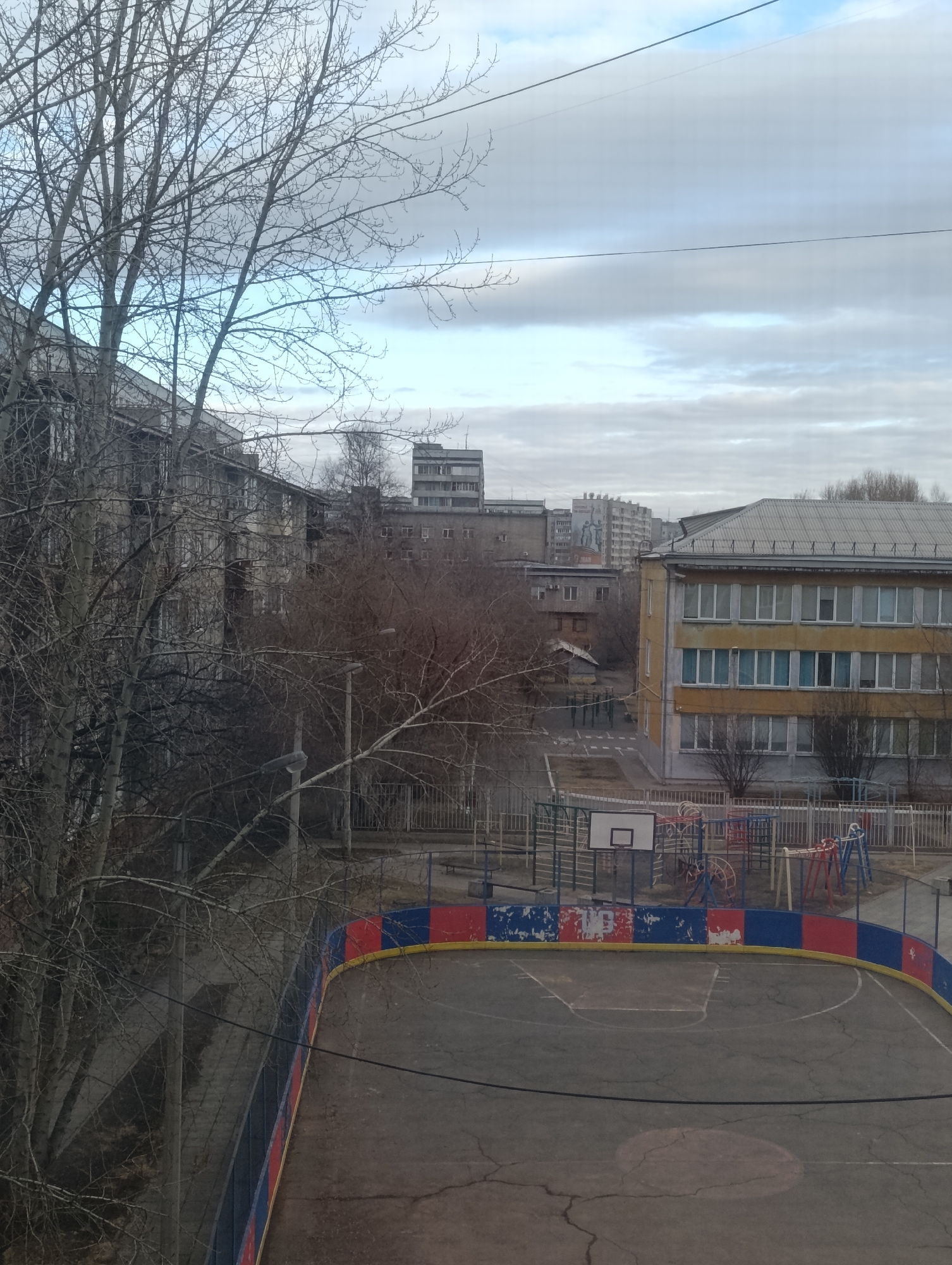 An elevated perspective shows an outdoor sports court with a low barrier wall of alternating red, yellow, and blue panels, situated in a courtyard within an urban setting. The court's surface is cracked and marked with faded white lines, and a basketball hoop stands at one end, next to a playground with swings and climbing equipment enclosed by metal fences. A large yellow and tan building with white-framed windows overlooks the court, along with gray apartment buildings and bare trees. The overcast sky creates a muted color palette throughout the scene, which has been determined to be in the city of Krasnoyarsk, Russia. There are no people visible in the image. Overhead power lines are also present.
